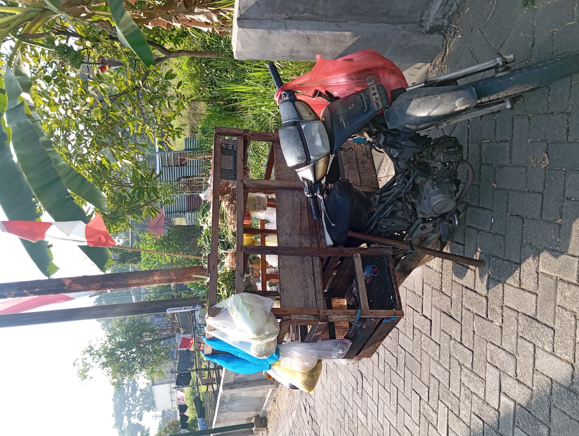 A motorcycle is parked on a brick sidewalk in Pandaan, Indonesia. The motorcycle is equipped with a wooden rack that extends behind the seat, carrying various items. In front of the rack, several plastic bags are hanging, containing what appear to be vegetables, including large cabbages. Behind the rack, more produce is visible. A red plastic bag is tied to the motorcycle's handlebars. The scene is outdoors, with trees and vegetation in the background, along with some corrugated metal structures. Two Indonesian flags are mounted on wooden poles near the motorcycle. The lighting suggests it is daytime, with sunlight casting shadows on the ground.