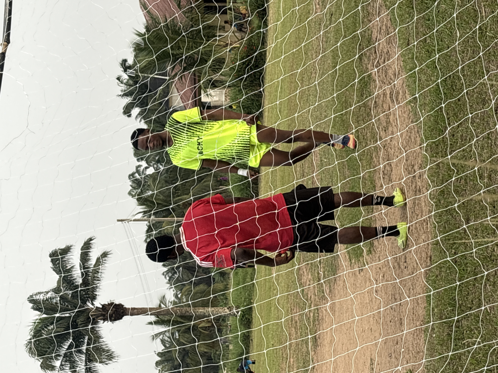 Two men stand on a field, possibly in Ikot Ekpene, Nigeria, as palm trees are visible in the background. The foreground is dominated by a white net, likely a soccer goal. One man wears a neon yellow and black jersey and matching shorts and is looking directly at the camera. The second man is closer to the camera, wearing a red shirt and black shorts. The field appears to be a mix of grass and bare earth.