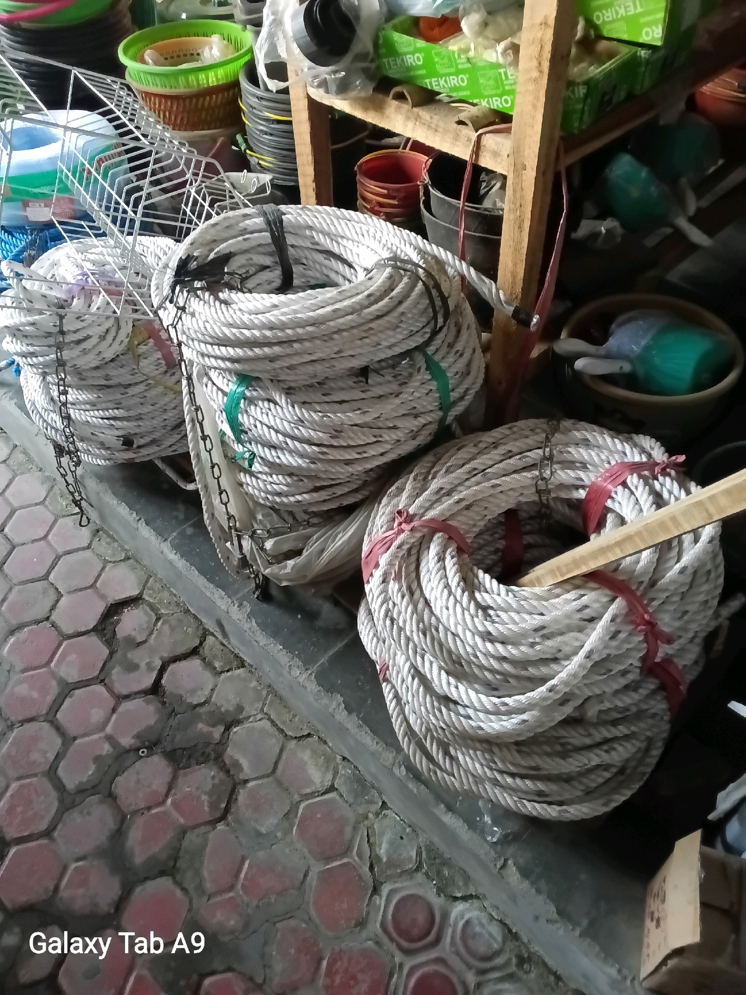 The image shows three coils of thick, white rope stacked on top of each other. The ropes are bound with green and red ties. In the background, there are shelves with various items, including plastic baskets, buckets, and what appear to be boxes of goods. The scene is set in what looks like a storage area or a shop. The floor is paved with hexagonal tiles. A "Galaxy Tab A9" watermark is visible in the bottom left corner of the image. There is no indication of the specific location in Ngulanwetan, Indonesia.