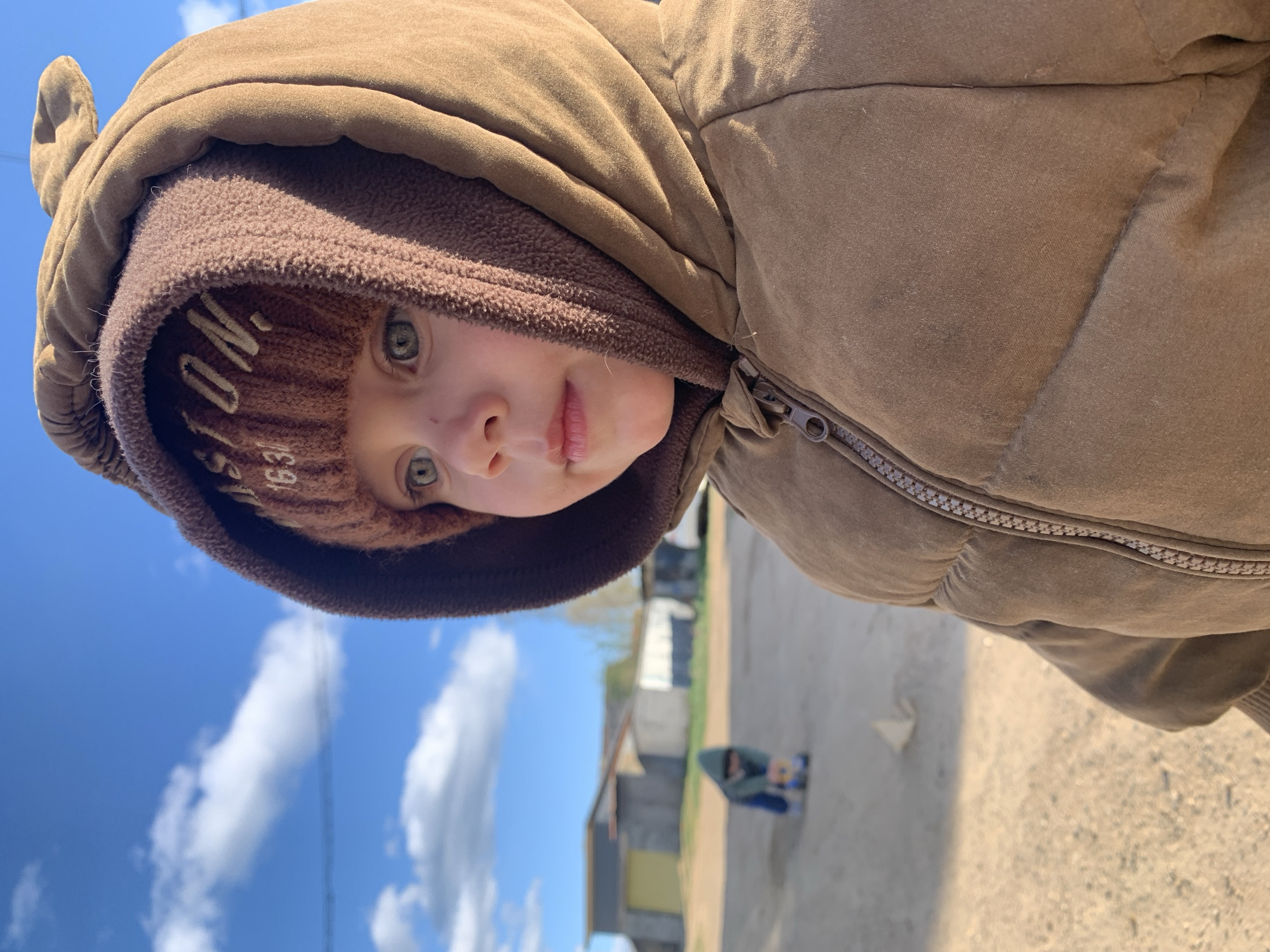 A young child with light eyes is partially obscured by the layered clothing. The child is wearing a brown knit cap with lettering, covered partially by a brown fleece hood and a brown puffy hooded jacket. The jacket is zipped to the upper chest. The view of the scene is angled down, showing a view of blue sky, a few puffy white clouds, buildings, a second person kneeling, and a rough, textured ground.
The geolocation given places this scene in Фоки, Russia.