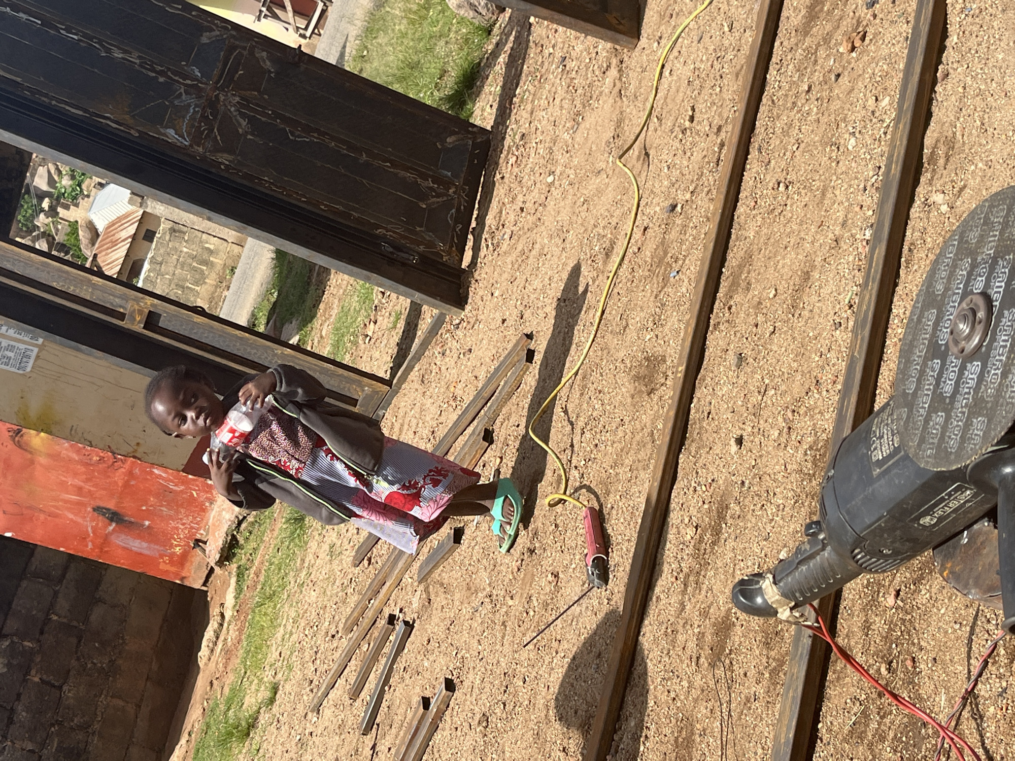 In Jos, Nigeria, a young child stands in a dusty, outdoor area surrounded by metal framing materials. The child, wearing a patterned dress and a dark jacket, holds a plastic bottle. To the child's left, a small red angle grinder with a yellow cord is visible on the ground, next to a metal rod. In the background, a large, weathered red metal sheet stands upright, and beyond that, a dark, heavy metal door frame is propped open, revealing a view of buildings and rocky terrain. The ground is covered in coarse sand and gravel, with patches of dry grass.