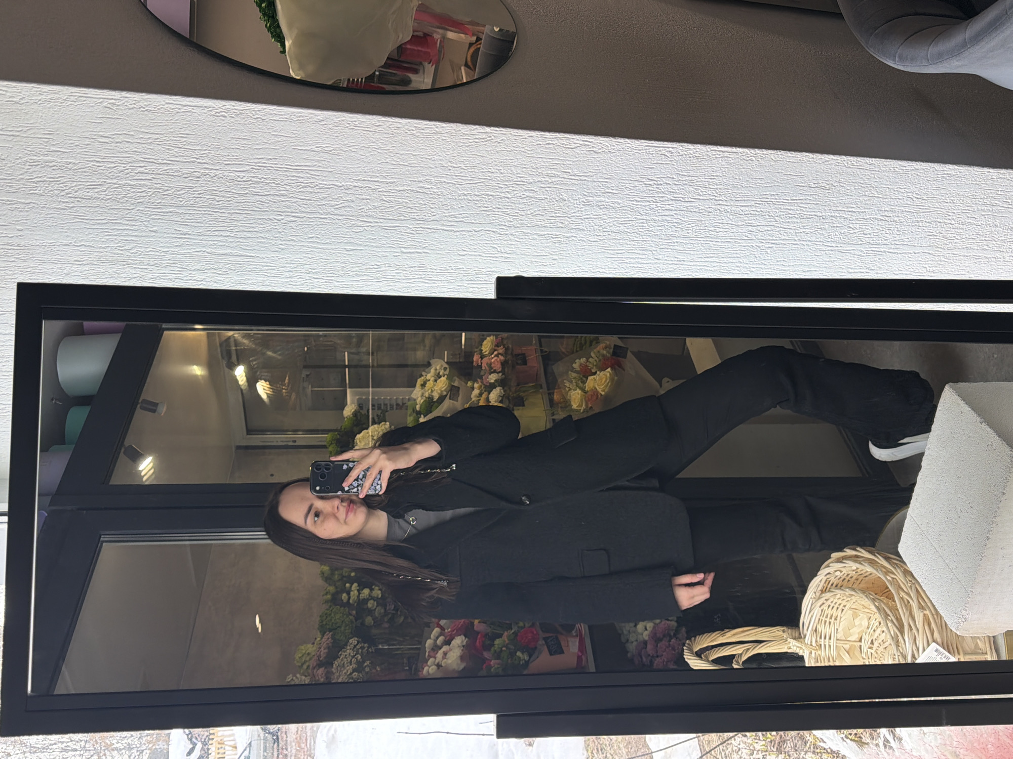 A young woman is depicted taking a selfie in a full-length mirror within a flower shop in Chaikovsky, Russia. The woman has long, dark hair, partially styled with braids on her left side, and is wearing a black blazer over a grey top with a heart-shaped pendant, black pants, and white sneakers. She holds a dark-colored smartphone with a patterned case in her right hand, angled to capture her reflection, and displays a slight smile.
The mirror's reflection reveals the interior of the flower shop, featuring numerous bouquets and floral arrangements in various colors (including pink, yellow, white, and purple) displayed on shelves and in cases behind the woman. Overhead artificial lighting is visible in the reflection. Through the shop's glass exterior, a blurred outdoor environment appears, indicating a cold setting with visible snow and bare trees.
In the immediate foreground, outside the mirror's reflection, three woven wicker baskets are stacked on the floor to the left, adjacent to a white rectangular concrete block. To the right of the mirror, a white textured wall is visible. A portion of an oval mirror with a black frame and a grey upholstered chair or similar furniture item are partially visible further to the right.