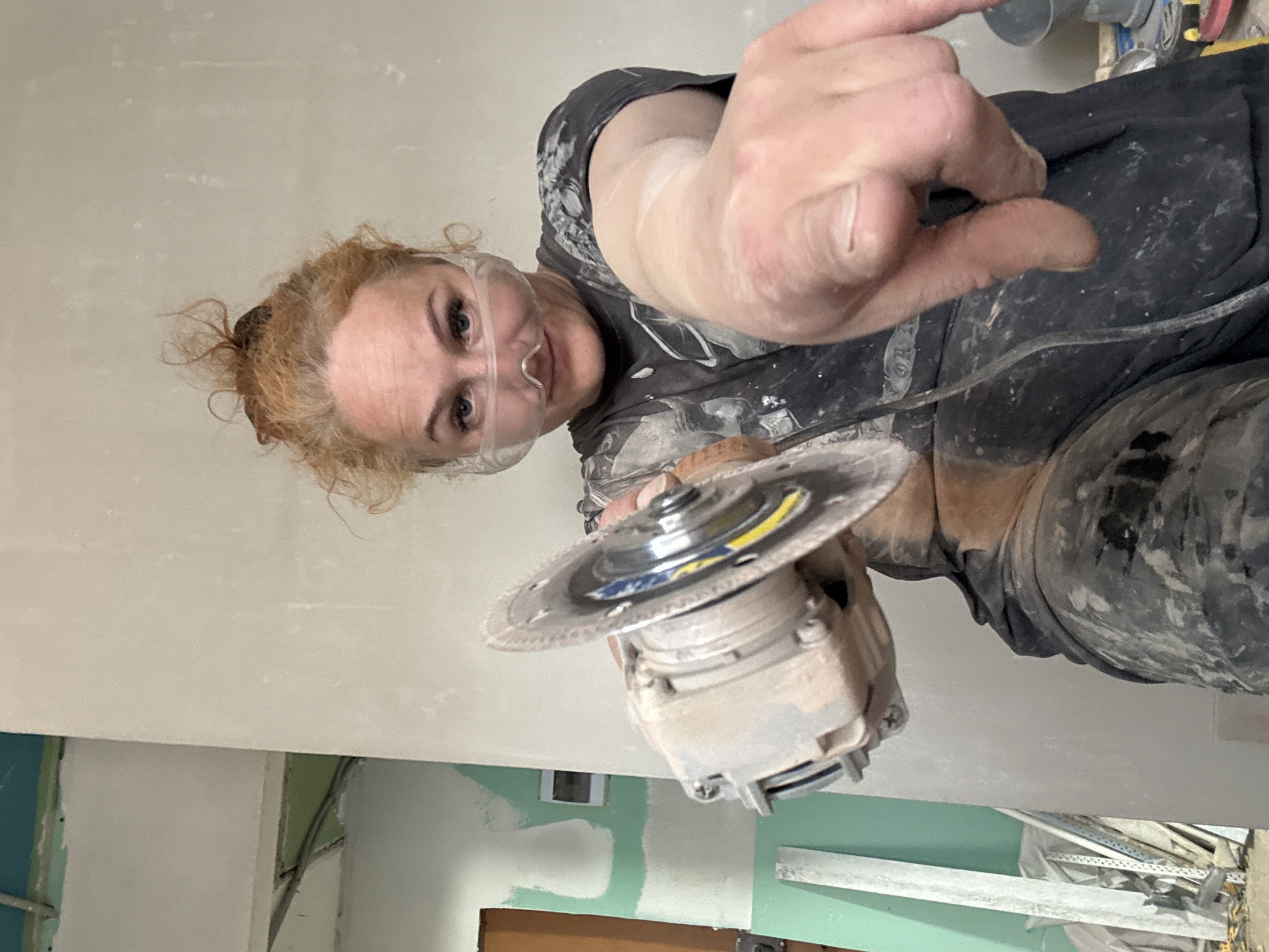 A woman is holding a power tool and pointing at the viewer. She has red hair pulled back in a bun, fair skin, and is wearing safety glasses. She is wearing a black shirt with white paint splatters. She holds a power tool with a silver circular blade. Her hands and face appear to have dust on them. The background is an unfinished interior with pale green and white walls. Construction materials and tools are visible near the floor. There is no definitive evidence to confirm or deny the location is Chaikovsky, Russia.
