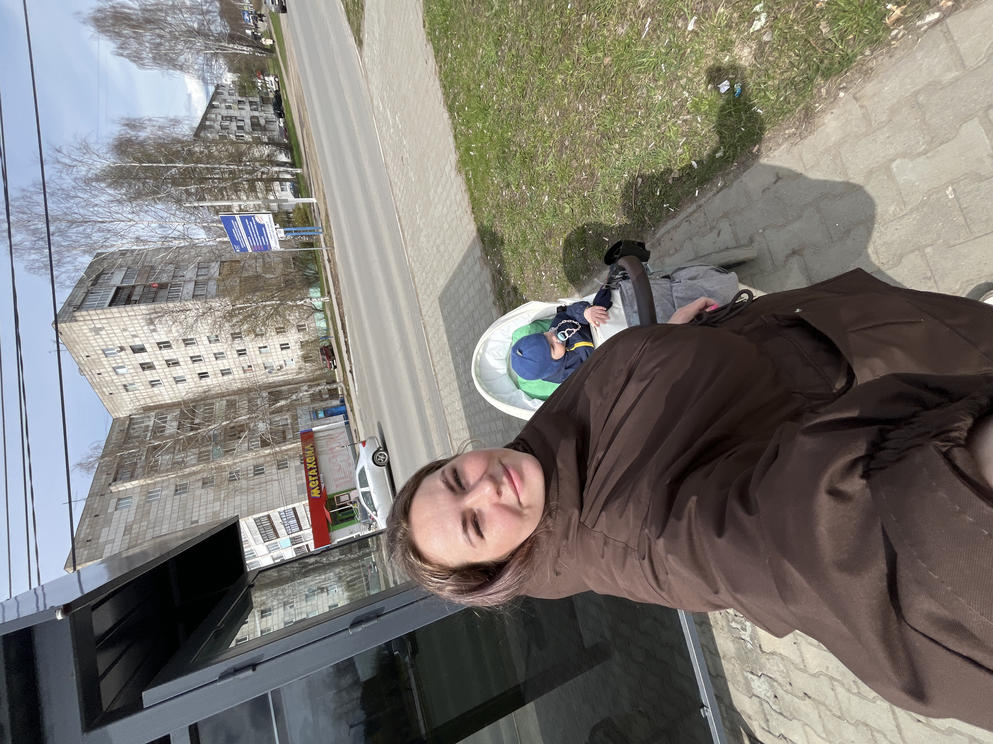 A woman is taking a selfie with a child in a stroller on a sidewalk in Chaikovsky, Russia. The woman is wearing a brown jacket and has short brown hair. The child is wearing a blue hat and jacket and is lying in a white stroller. In the background, there is a street, a grass field, and an apartment building. There is also a storefront labeled "MEГAXEHA".