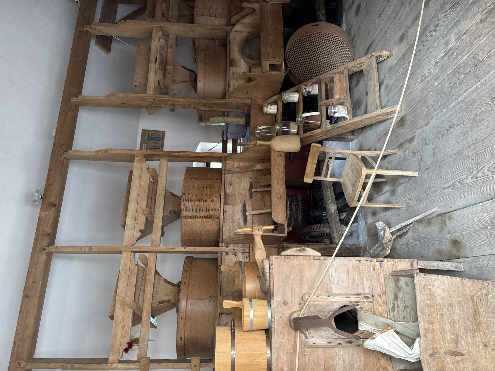 The image depicts the interior of a historical mill, likely part of the Postojna Cave system in Postojna, Slovenia, given the context of its operation as a tourist attraction. The scene is dominated by aged wooden machinery and equipment, indicative of traditional grain processing. Prominently featured are large wooden bins and hoppers, designed to hold and channel grain. Several wooden buckets of various sizes are arranged on a lower platform, some with metal bands. A wooden chair sits in the foreground, positioned on a weathered wooden floor. A set of wooden stairs leads up to a collection of glass jars filled with granular substances, presumably flour, grains, or spices, suggesting a functional display of the mill's ingredients or products. Ropes are strung across the foreground, possibly to demarcate visitor pathways. The overall environment is rustic, with a focus on the historical functionality of the mill.