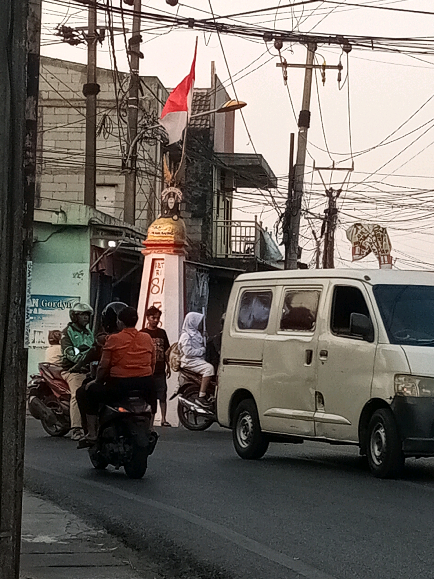 In Karangsatria, Indonesia, a street scene unfolds with a white van driving on the right side of the road. To the left, three people on two motorcycles are present. One motorcycle has two riders, a person in an orange shirt in the back and another in a green jacket in the front, wearing a helmet. The second motorcycle is also occupied by a person wearing a white headscarf. A pole with electrical wires and a red and white Indonesian flag is visible in the background, alongside a building with a brick facade. The weather appears overcast.