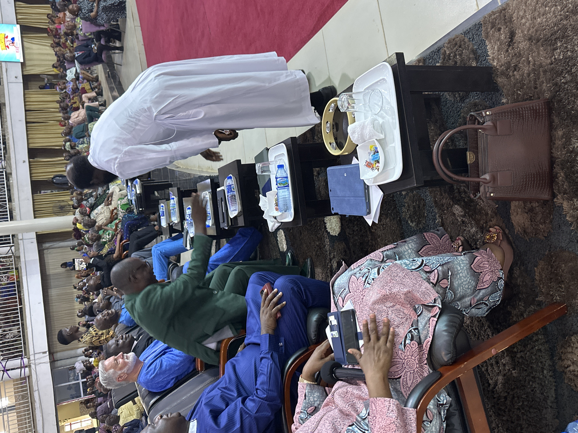 The image depicts an indoor gathering, likely a religious service, taking place in Television, Nigeria. A group of people are seated in chairs, many facing forward towards an unseen focal point. In the foreground, a man in a white robe is bent over, interacting with a small table. On the table, there are several items including a tambourine, a white towel, glasses, a small dish with candies, and a tablet. To the left of this man, a woman in a patterned pink and gray dress is seated, holding a tablet and a microphone. Another person in a blue outfit is seated to her left. In the background, more individuals are seated, some looking towards the front, while others are engaged in conversation. The environment appears to be a large hall with cushioned seating and a red carpet visible in the background.