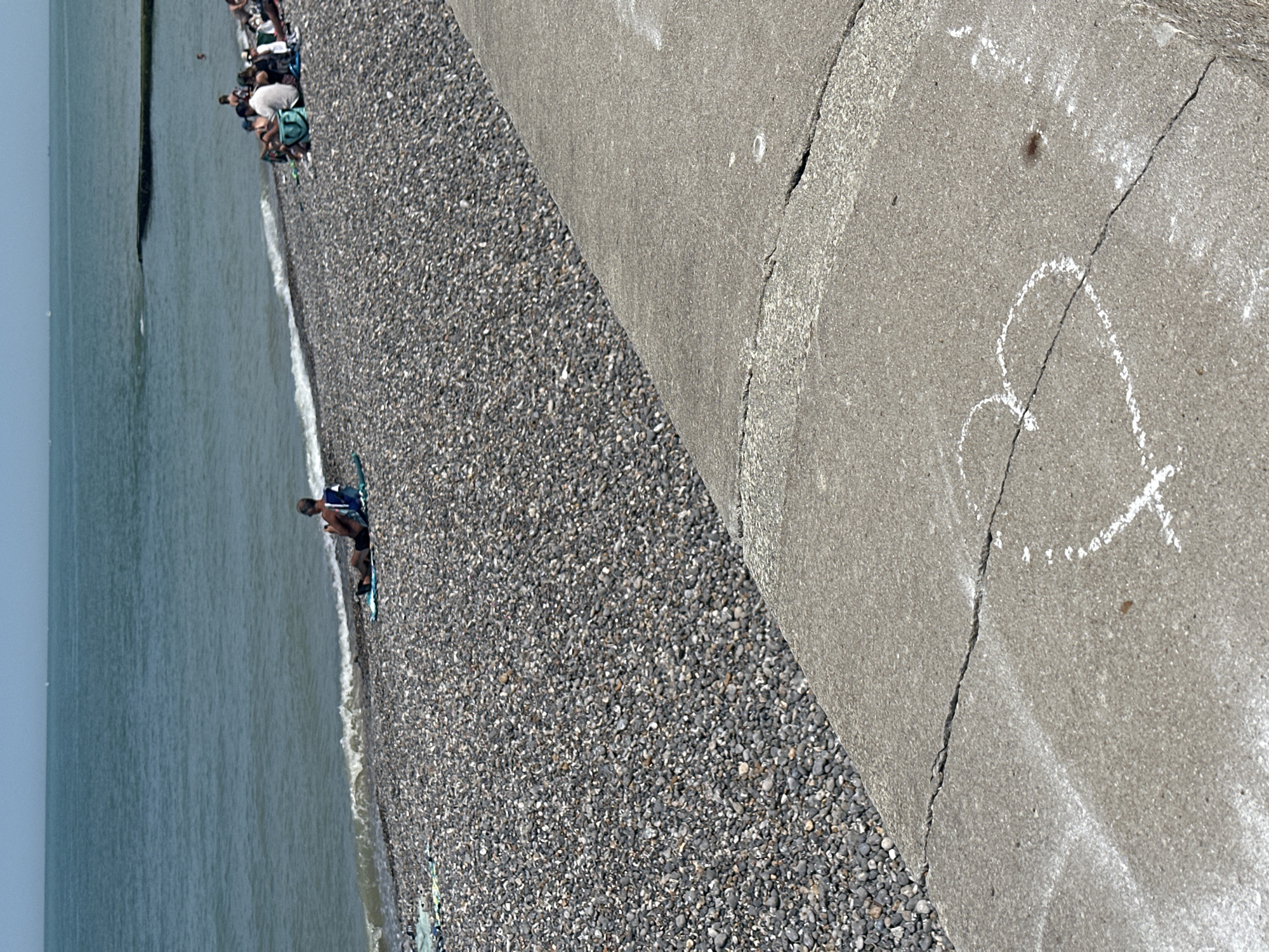 A long shot captures a sunny day at a pebble beach in Hautot-sur-Mer, France. In the foreground, a concrete walkway with visible cracks features a heart drawn in chalk. Beyond the walkway, the pebble beach is populated with several groups of people relaxing. Further out, the calm, blue sea stretches to the horizon under a clear sky. A man sits alone on a towel by the water's edge, facing the sea. Several other people are seated on towels and in chairs on the beach, some appearing to be in conversation. The scene is devoid of any dramatic action, presenting a peaceful, everyday beach setting.