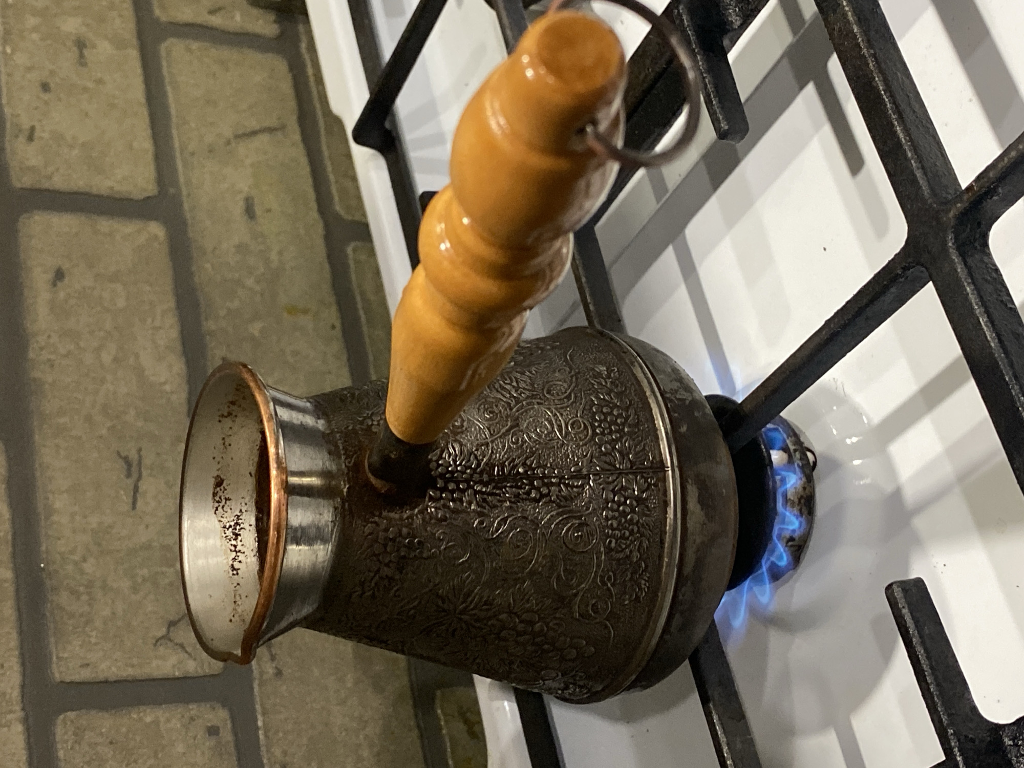 This image depicts a metal Cezve, also known as a Turk or Ibrik, containing coffee, positioned on a white gas stove with black cast-iron grates. The Cezve, dark in color, features an embossed pattern of grapevines and leaves, and has a light brown, ridged wooden handle extending to the right with a metal hanging ring at its end. The top interior rim of the Cezve shows dark coffee residue. Beneath the Cezve, a gas burner emits blue flames, indicating active heating. The background consists of a wall covered with light-toned, rectangular brick-patterned tiles with dark grout lines. The scene is located within a kitchen setting in Sochi, Russia. No individuals are present.