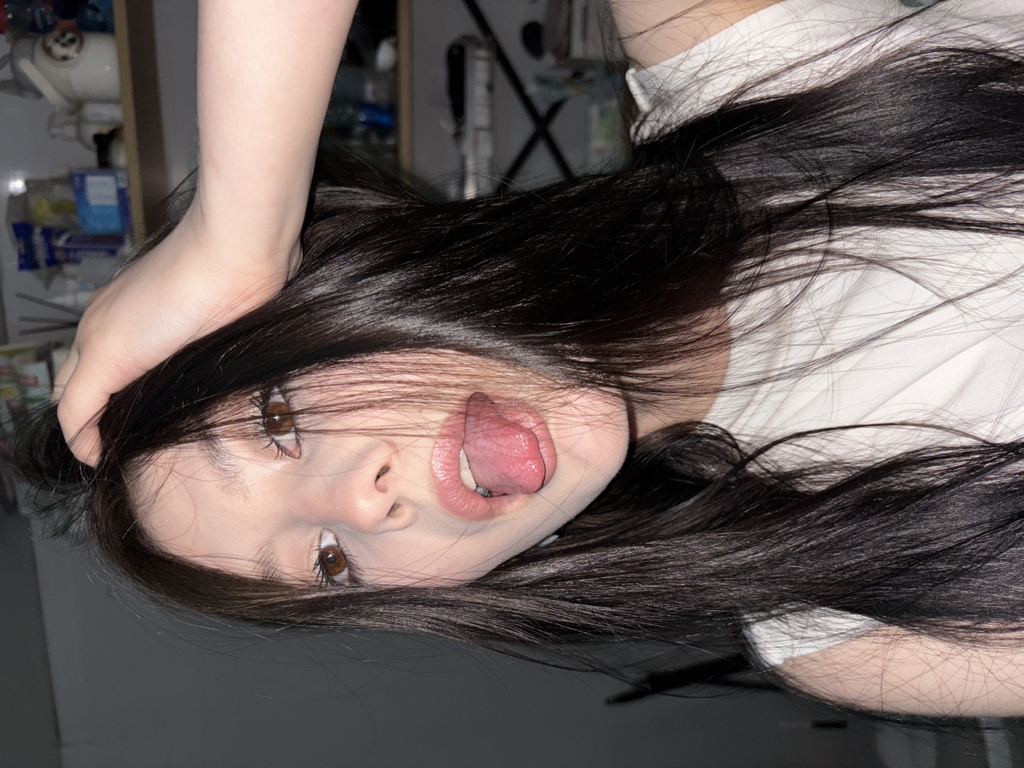 The image captures a close-up, rotated portrait of a person, likely female, with long, dark hair. The subject's head is tilted significantly, with her face oriented towards the camera. She has light skin, dark brown eyes, and is making an expressive face with her tongue partially extended. Her lips appear to be slightly parted, revealing some teeth and her tongue.
Her dark, flowing hair is prominent, framing her face and spreading across the frame, suggesting movement or a dynamic pose. A hand and forearm are visible in the upper left, appearing to be holding or resting on her head or hair. She is wearing a light-colored, possibly white, top, visible around her shoulder and arm.
The background is dimly lit and out of focus, showing blurred shapes that suggest an indoor setting, possibly shelves or furniture with indistinct items. There are no clear identifying objects or text in the background. The overall impression is playful and expressive. The image is taken indoors, and there are no specific visual cues to indicate the stated location of Фоки, Russia.