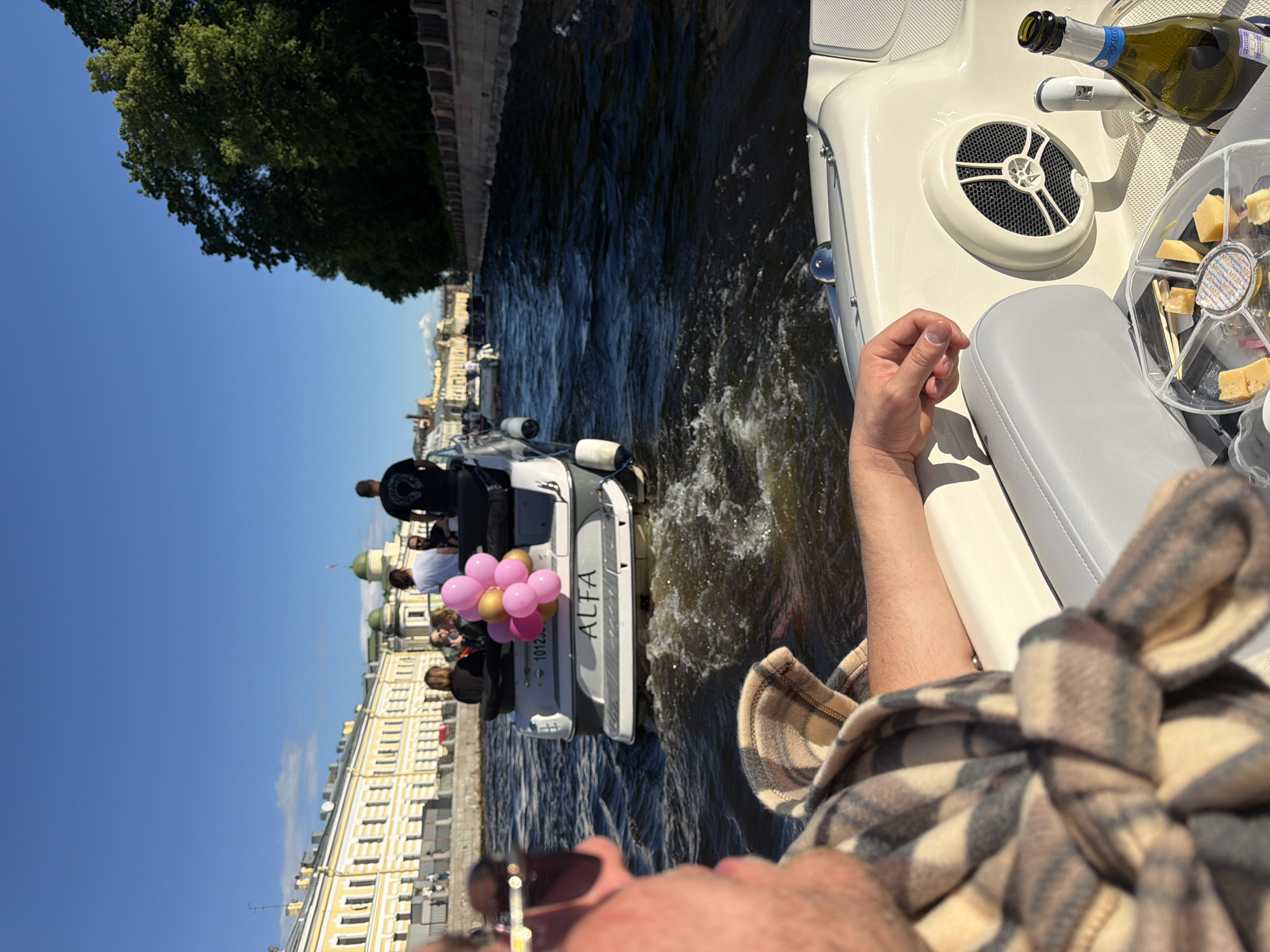 This image captures a sunny day on a waterway in an urban setting, possibly a canal or river. The perspective is from a person in a boat, looking forward.
In the foreground, the arm and shoulder of the person taking the photo are visible, draped in a plaid blanket. To the right, a part of the boat's interior is seen, featuring a gray seat, a circular speaker, a bottle of Prosecco, and a clear plastic tray with cheese cubes and skewers.
Further ahead, another boat is moving away from the camera. This boat has several passengers, including individuals wearing white and black shirts, and a person with blonde hair. The back of this boat displays the name "ALFA" and a number "1012". A cluster of pink and gold balloons is attached to the rear of this boat.
The waterway is flanked by buildings on the left, appearing to be historical architecture with yellow facades and numerous windows. On the right, a tree-lined embankment is visible, with lush green foliage. The sky is a clear, bright blue. The water is choppy, indicating the movement of the boats. The overall scene suggests a leisurely outing on the water.