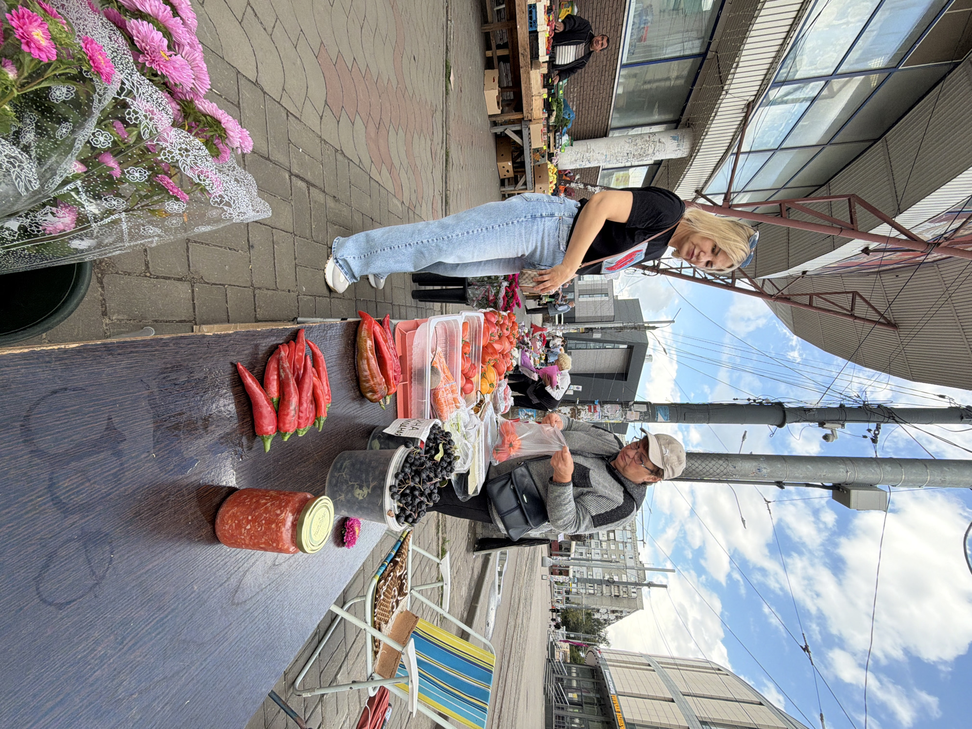 A blonde woman in a black t-shirt and light blue jeans stands at a market stall, looking towards the camera. To her right, an older person wearing a cap and a grey and black striped jacket is bagging groceries. In the foreground, a wooden table displays red peppers, a jar of red paste, and a bucket of dark berries. Behind the table, a striped folding chair sits empty. In the background, a man in a dark jacket stands at another stall piled with produce. The market appears to be located on a paved street in a city, with buildings and overhead wires visible. This scene is likely taking place in Nizhny Novgorod, Russia, given the context provided.