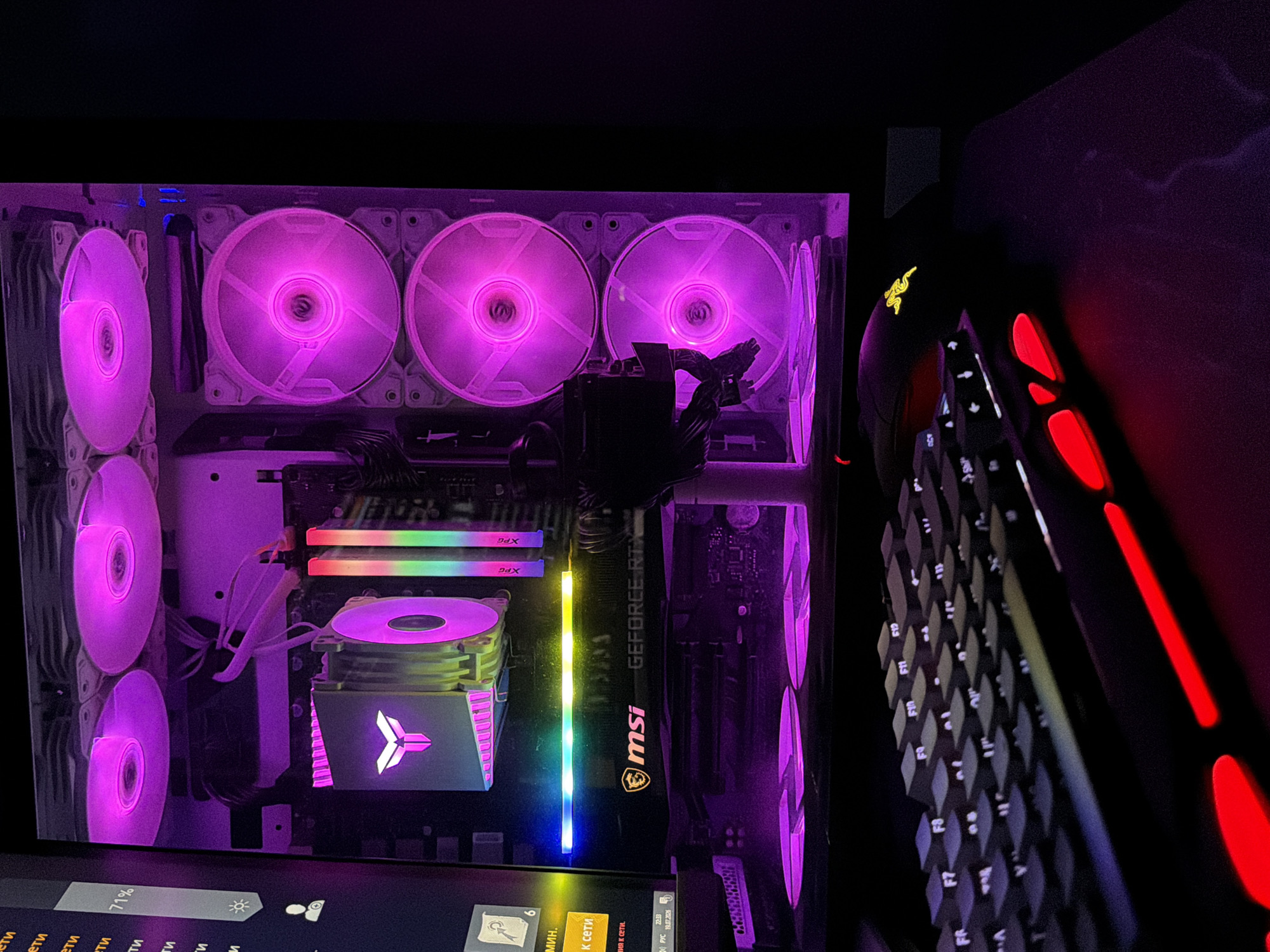 The image shows the interior of a computer tower and a keyboard. The computer has multiple fans with pink lighting, RGB RAM sticks, and an MSI GeForce RTX graphics card. The keyboard is black with illuminated red accents and white keycaps. A Razer mouse is partially visible next to the keyboard. The scene is dimly lit with a pink and purple ambient glow from the computer's lighting.