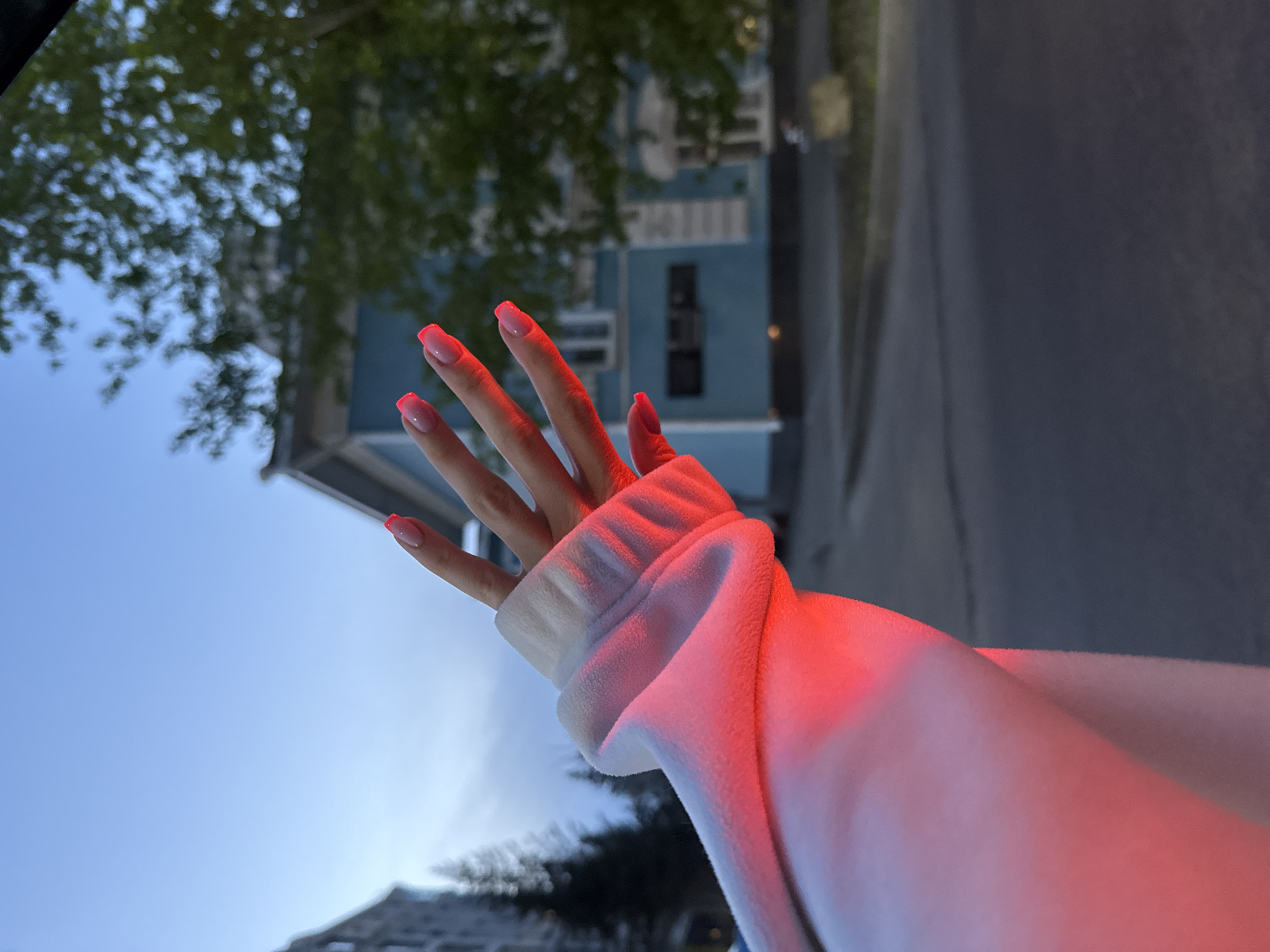 A low-angle, full-frame photograph shows a person's left hand and arm raised against a background of a partially obscured building and an overcast sky. The building, likely a residential structure, is painted light blue and appears to have multiple windows and architectural details. The right portion of the scene shows a section of a roadway or sidewalk. The subject's hand is palm forward, with fingers extended. The fingernails are painted with a light pink color. The person wears a white long-sleeved garment with a cuffed wrist, the sleeve is catching a reflection and is colored in red. The background sky is overcast, with bare tree branches visible in the periphery.
