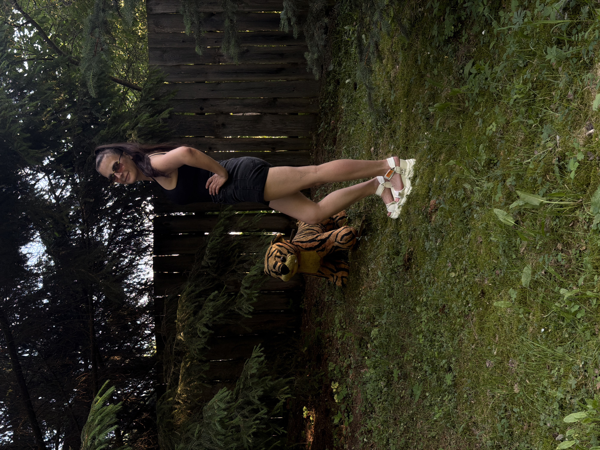 In a yard in Krasny Mayak, Russia, a woman poses with her hand on her hip. She is wearing a black tank top, black denim shorts, and white platform sandals. To her left, a stuffed tiger sits on the grass. The background consists of a wooden fence and trees. The ground is covered in grass and some small plants.