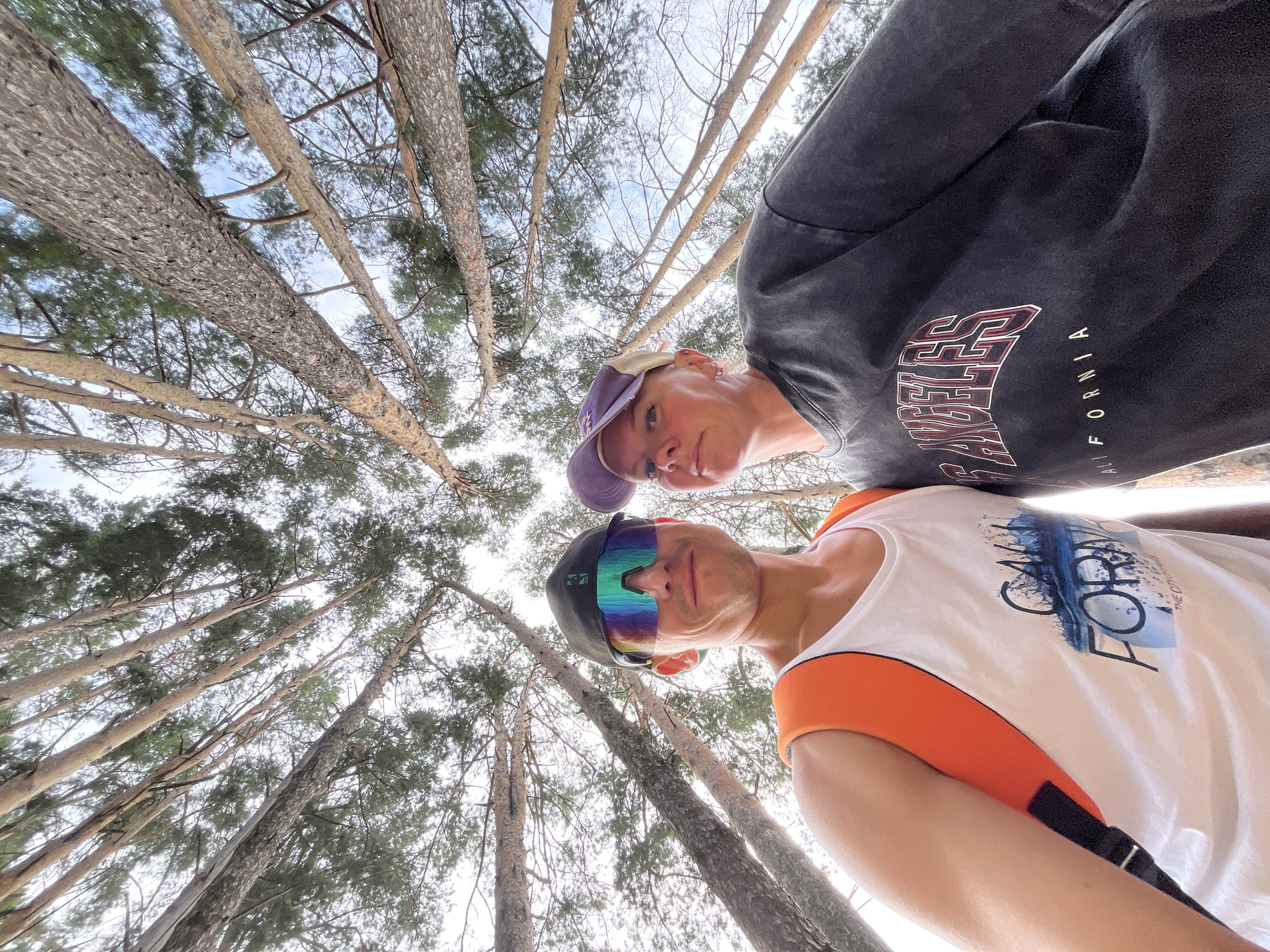 A low-angle, wide shot captures a man and a woman posing for a selfie amidst tall trees in Chaikovsky, Russia. The man, on the left, wears a white tank top with an orange backpack and futuristic sunglasses. The woman, on the right, is dressed in a dark, oversized t-shirt and a purple baseball cap. The background is dominated by the trunks and branches of pine trees, reaching towards a bright, overcast sky. The man's tank top has a graphic print that includes the words "CALL FORME" and "THE CITY OF DREAMERS." The woman's t-shirt features the text "LOS ANGELES CALIFORNIA." The setting appears to be an outdoor, forested area within the city limits of Chaikovsky.