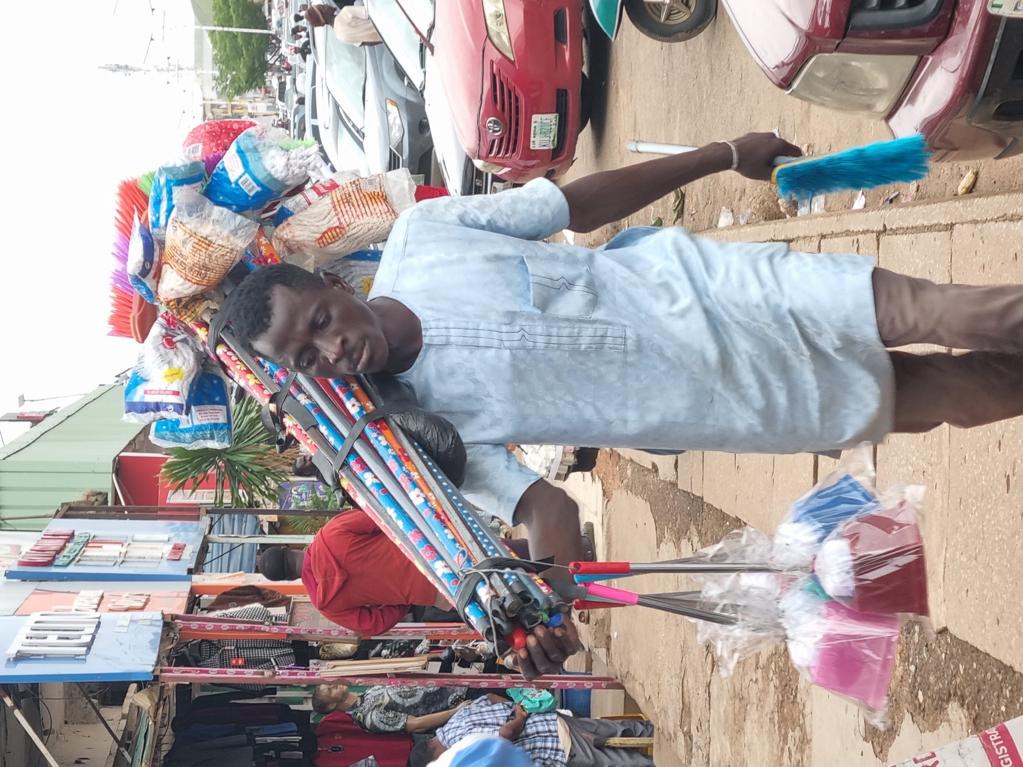 In the city of Jos, Nigeria, a Black man in a light blue, long-sleeved garment with a breast pocket walks along a sidewalk carrying a large bundle of cleaning supplies, primarily brooms and mops, on his left shoulder. He appears to be a street vendor. The brooms have colorful patterned handles and bright bristles, while the mops are individually bagged in clear plastic with colored tops. He is also holding a blue feather duster in his right hand.
The background reveals a busy street scene. To the left, there is a row of market stalls with clothing displayed. Further back, there are more stalls and shops, some with signage. Vehicles are visible on the road, including a red car with a license plate that reads "KUJ 821CF". The ground is paved with concrete. The overall atmosphere suggests an active marketplace in a Nigerian urban environment.