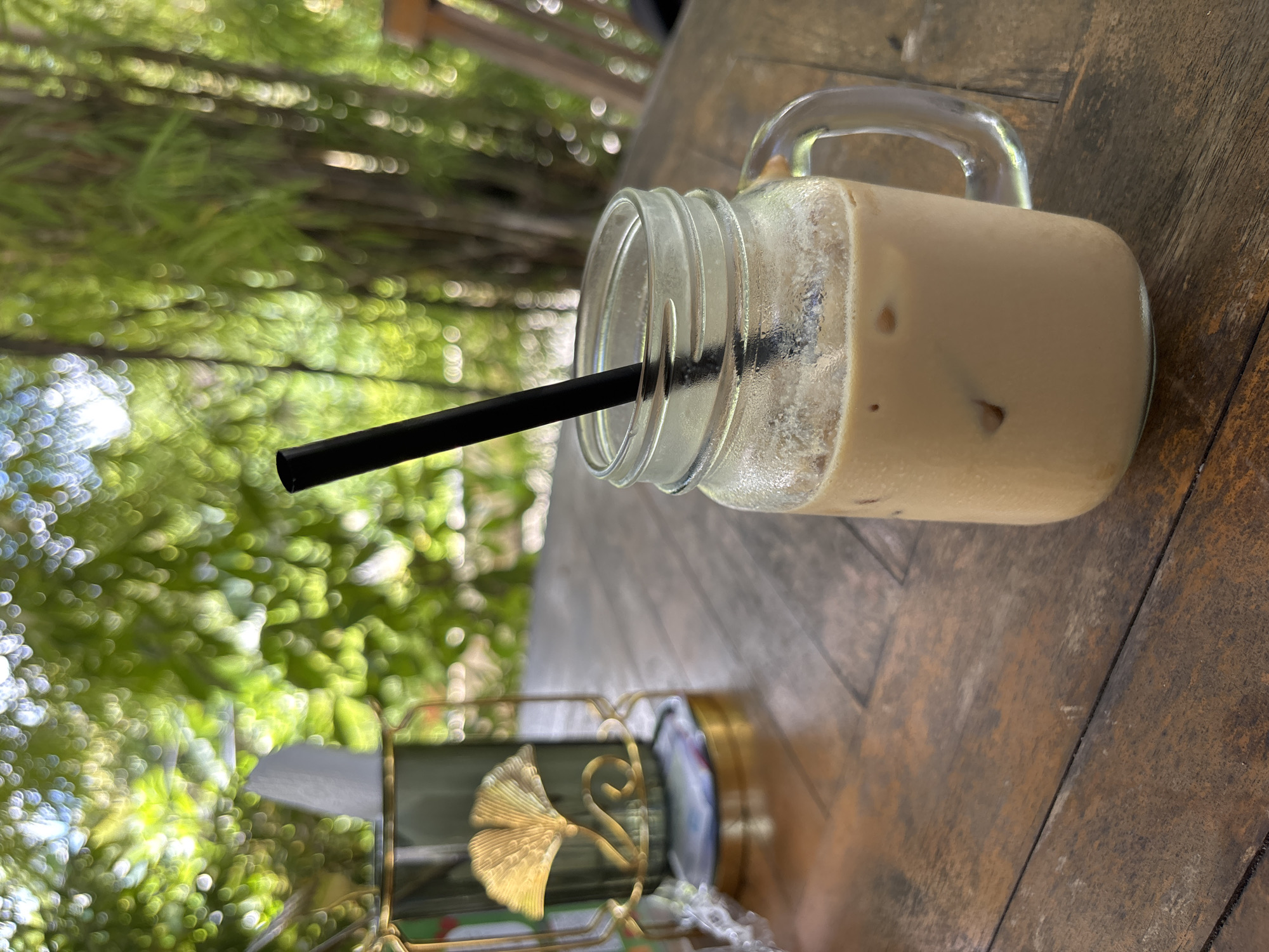 The image shows a glass jar with a handle, filled with a tan-colored drink and ice, on a weathered wooden surface. A black straw is inserted into the drink. Next to the jar is a tissue holder with a gold ornamental design. In the blurred background, greenery suggests an outdoor setting. The location is Ungasan, Indonesia, based on geographical inferences from the scenery.