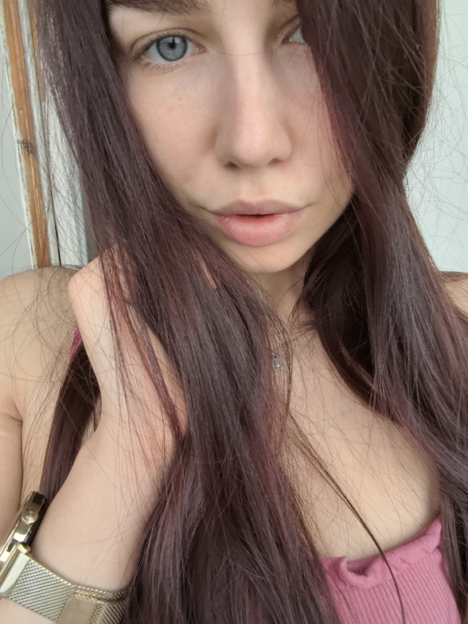 A close-up, self-portrait of a young woman with fair skin, long, dark auburn hair, and a singular blue eye visible. She is wearing a pink tank top with a ruffled neckline. A gold wristwatch with a mesh band is visible on her left wrist. The background is a plain, light wall. The image lacks any identifiable landmarks, making it impossible to confirm a location in Stara Zagora, Bulgaria. There are no discernible actions or interactions in the image.
