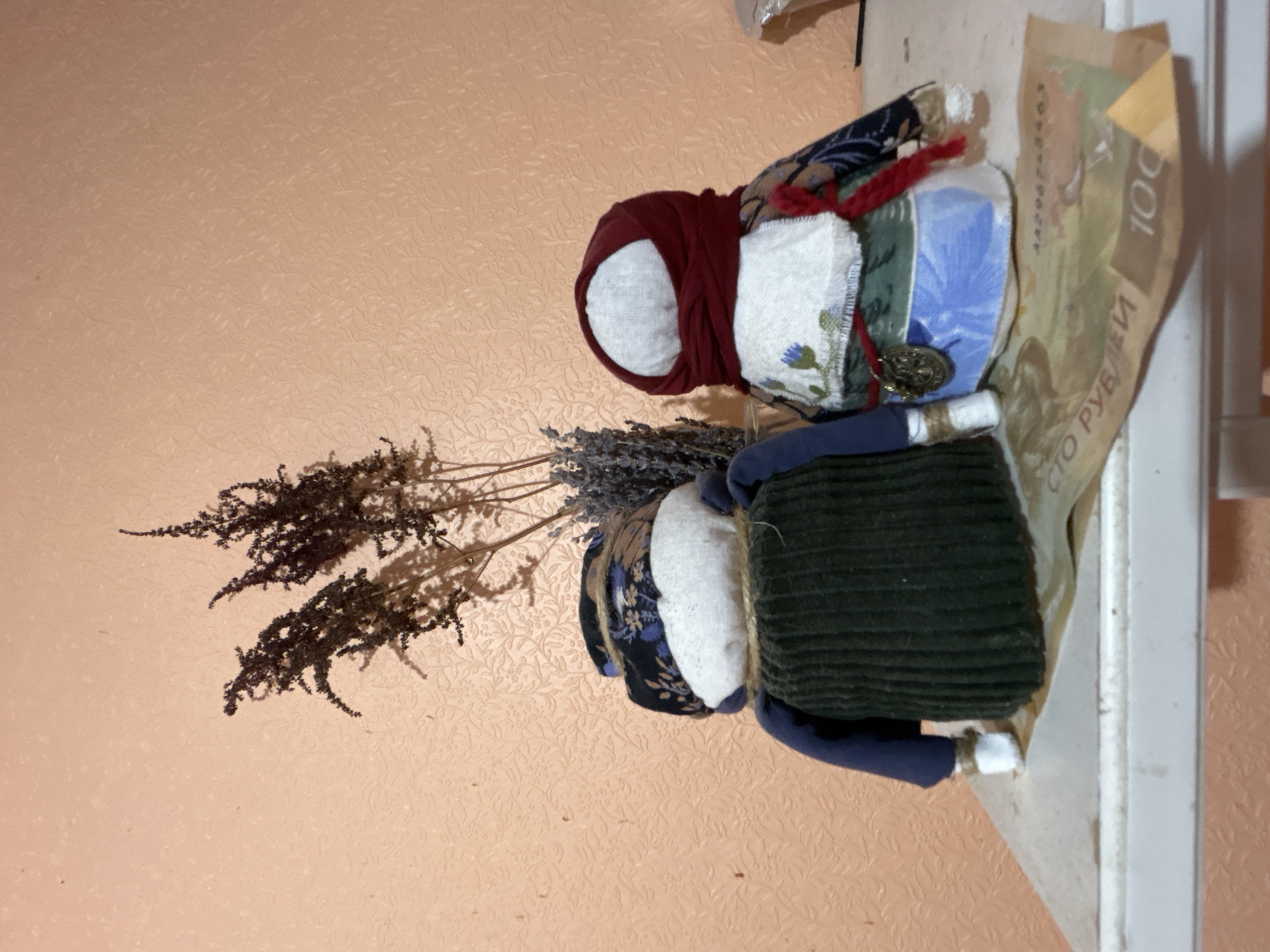 The image shows two handmade dolls, possibly folk art, positioned on a white shelf. The doll on the left wears a dark green corduroy dress over a blue garment and a patterned headscarf. The doll on the right has a red headscarf, a white apron with floral embroidery, and a blue skirt, adorned with a metal medallion. Behind the dolls, a dried floral arrangement, possibly lavender and other grasses, stands in a vase. A banknote, appearing to be a 100 ruble bill, is placed in front of the dolls on the shelf, partially obscuring the lower portion of their bodies. The background is a textured peach-colored wall. The scene suggests an indoor setting, potentially a home environment, with no indication of a specific location within Moscow, Russia.