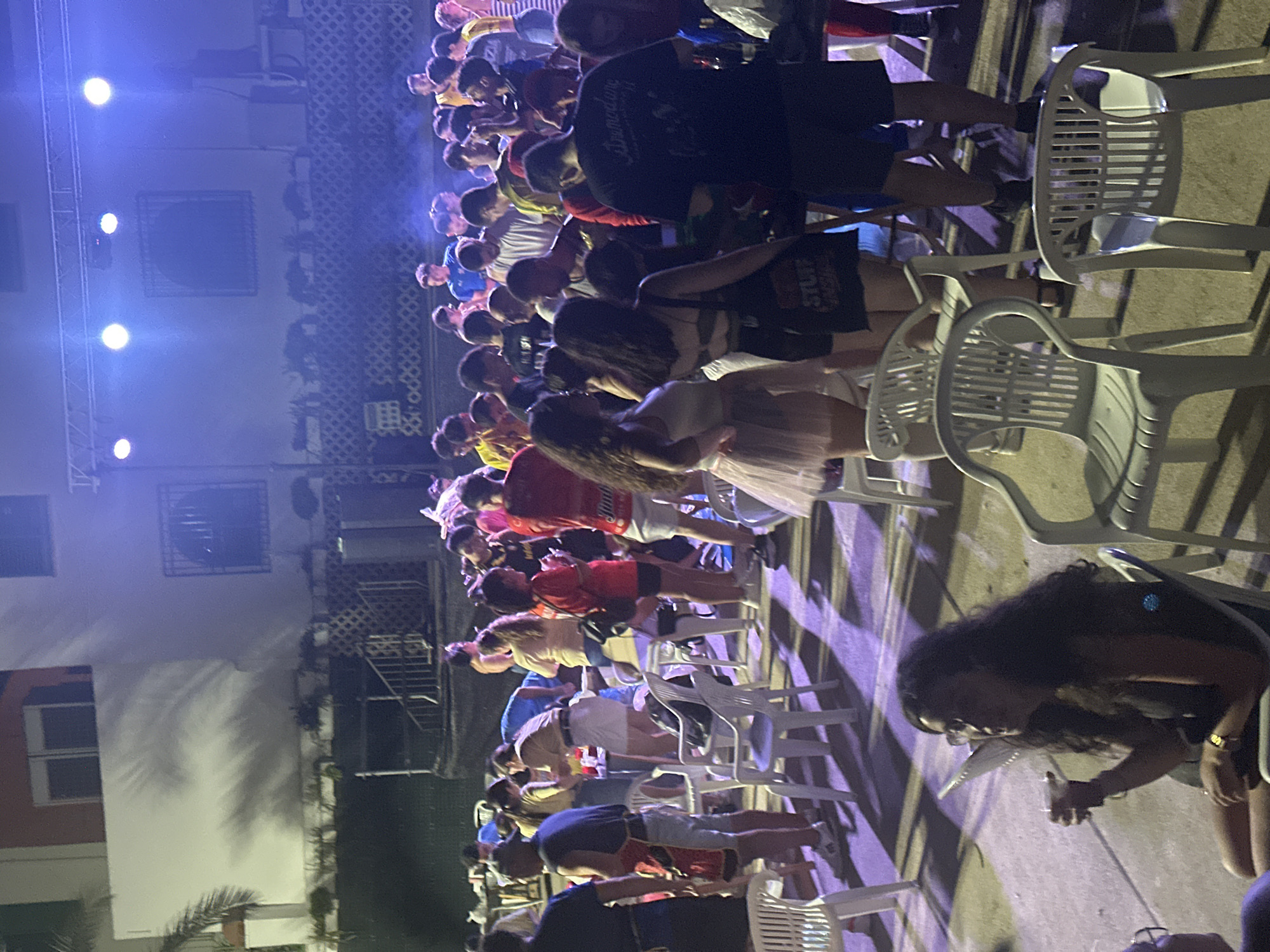 The image depicts an outdoor gathering in Blanca, Spain, possibly a festival or party, illuminated by stage lights. A crowd of young adults is visible, some standing and some seated on plastic chairs. In the foreground, a woman with long, dark curly hair is seated, holding a drink and looking to her right. She is wearing glasses and a dark sleeveless top, with a gold watch on her left wrist. Behind her, other individuals are present, and further back, a stage setup with lighting equipment is visible. The environment appears to be an open area with some architectural elements in the background, including buildings and railings. The overall atmosphere suggests a lively event taking place in the evening or night.