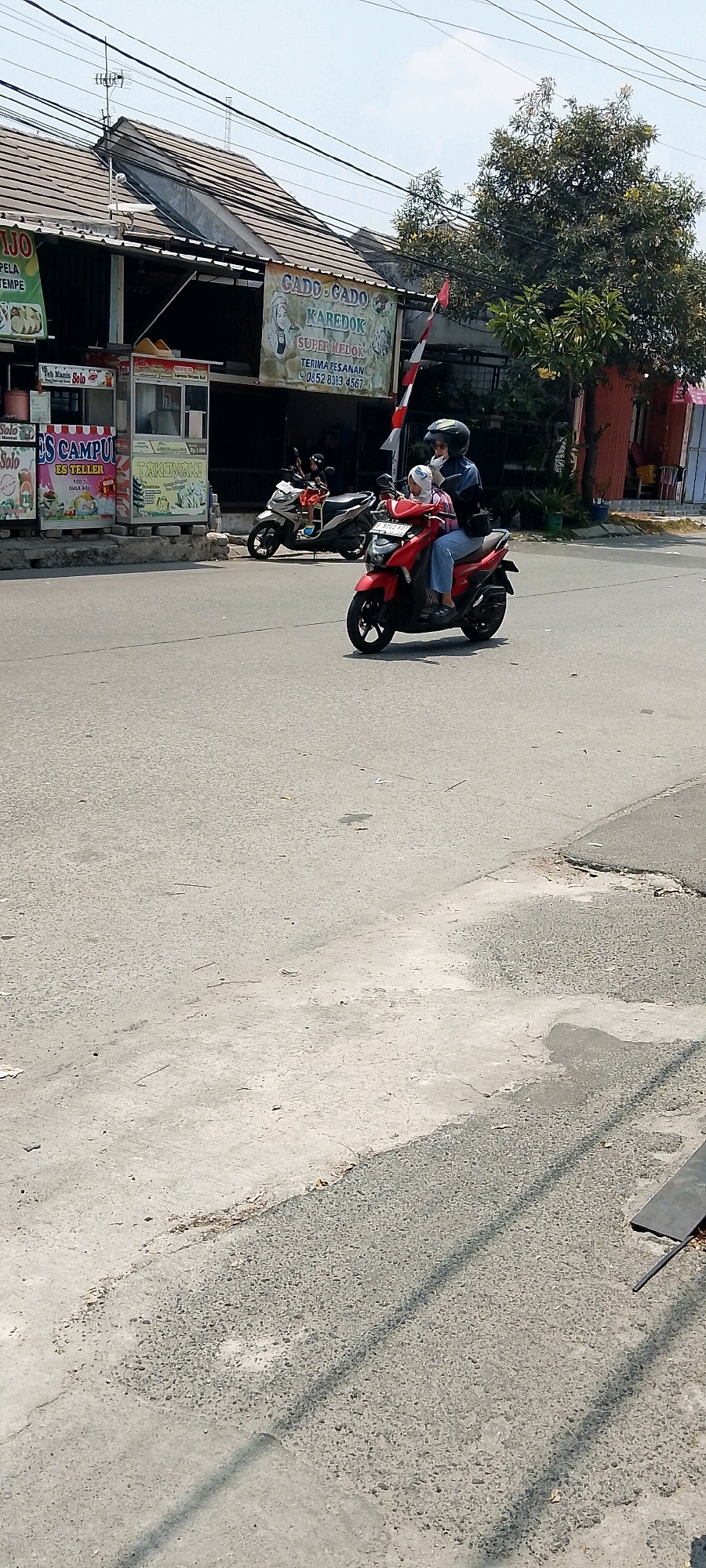 A red scooter is being ridden by a person wearing a helmet and blue shirt. A smaller person, likely a child, is sitting in front of the rider, also wearing a helmet. The scooter is on a paved road. In the background, there are buildings with signs advertising food items such as "CADO-CADO" and "TAKOYAKI." Another scooter is parked behind the red one. Trees and power lines are visible above the buildings. The sky is bright and clear. The scene appears to be in an urban environment in Satriamekar, Indonesia.