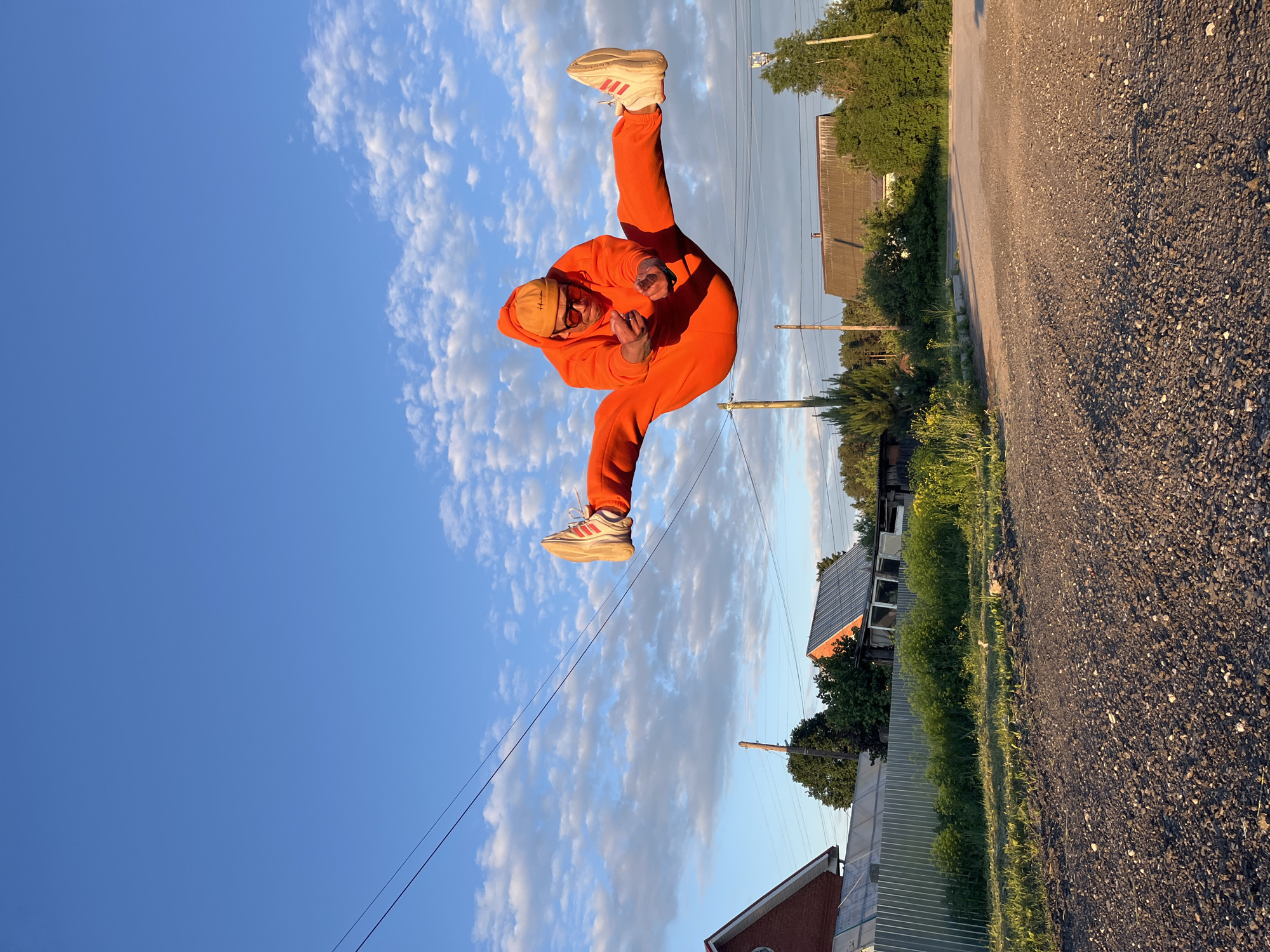 A full-body outdoor shot captures a person mid-air against a bright blue sky with scattered white clouds. The subject, a male, is dressed in a vibrant orange tracksuit, including a hoodie and sweatpants, light-colored sneakers with red stripes, and a tan beanie. He is in a dynamic pose, with his arms bent and one leg raised, looking directly at the camera.
The setting appears to be a rural or semi-rural environment, possibly in Chaikovsky, Russia. A gravel road runs along the right side of the frame, showing elongated shadows indicative of either late afternoon or early morning (golden hour). In the background, there are green trees, bushes, and some simple buildings with dark roofs. Utility poles and power lines are visible, stretching across the scene. The overall weather is clear and sunny. The person's playful pose and energetic stance are notable. No specific text is clearly legible on his clothing or accessories, though the shoes feature a three-stripe design.
