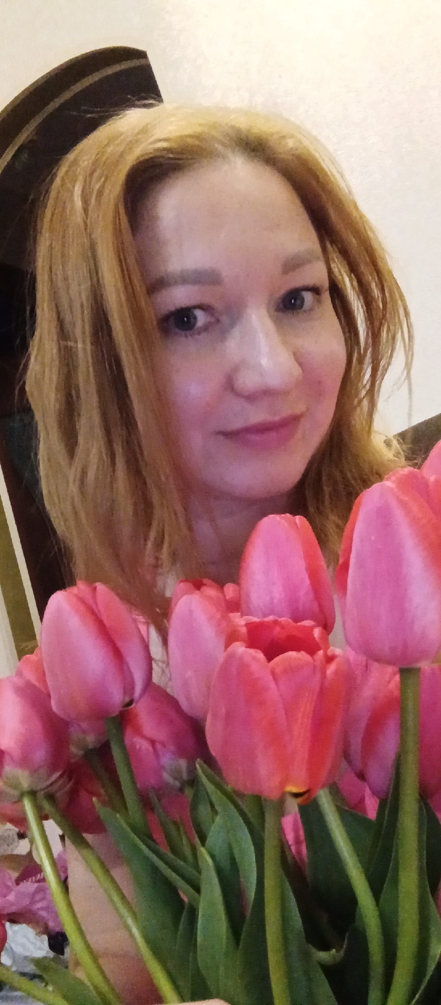 A medium shot depicts a woman with reddish-blonde hair holding a bouquet of pink tulips. She is fair-skinned with light blue eyes and a gentle smile. A dark-colored door frame is partially visible in the background on the left. The location is Unknown, Unknown.
