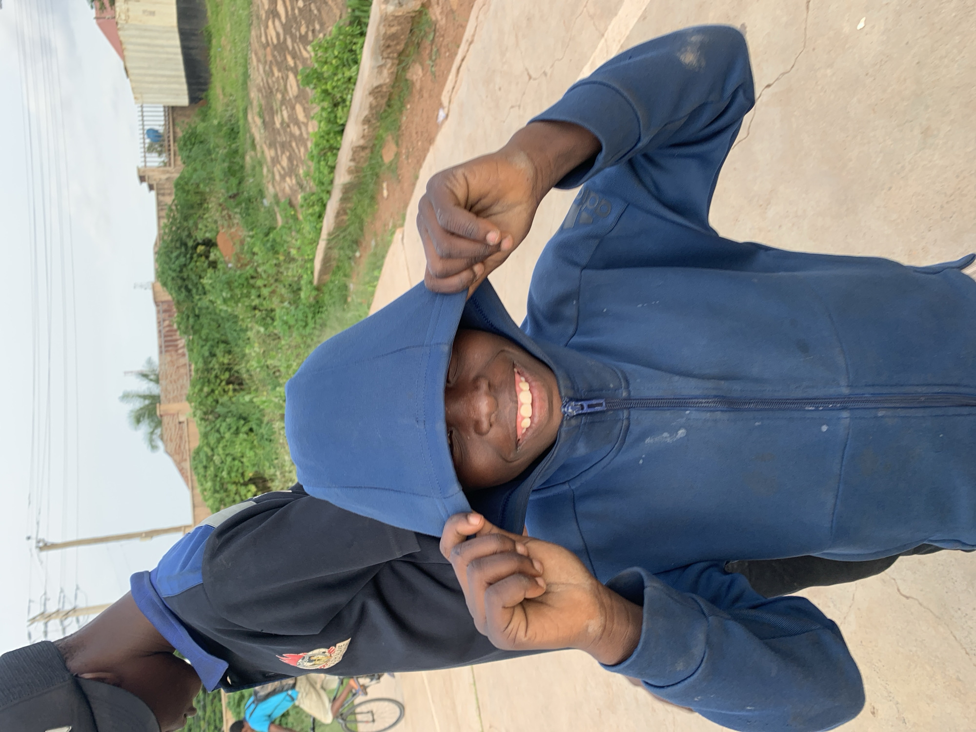 In the city of Gura Top, Nigeria, a young boy with a wide smile is pictured outdoors, holding the hood of his blue zip-up sweatshirt with both hands. He is wearing a blue hoodie and the fabric appears worn and slightly stained. Behind him, another person, partially visible, is wearing a black long-sleeved shirt with a blue collar and a black baseball cap. The background shows a dirt path with some greenery and buildings in the distance. The sky is overcast.