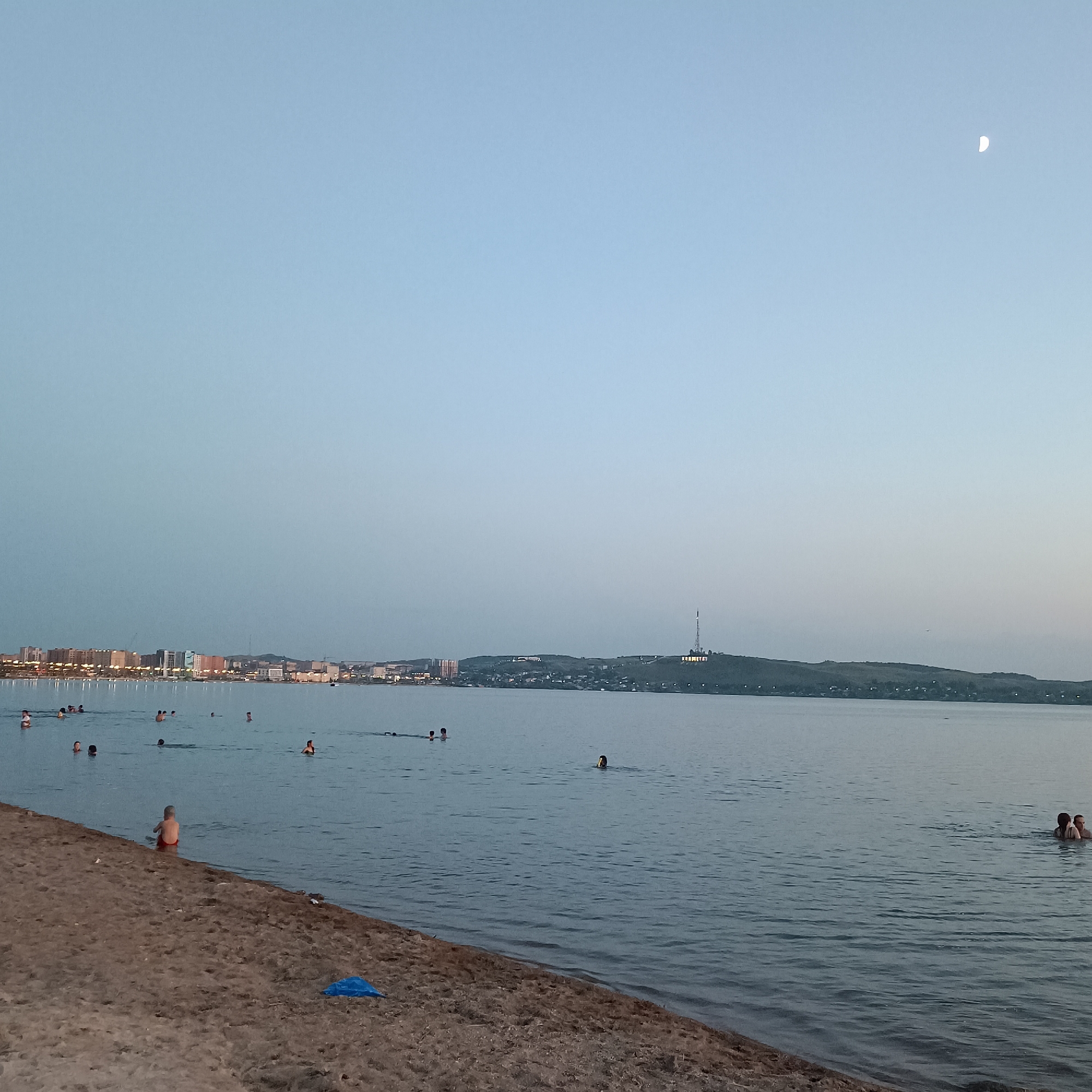 The image depicts a view of the Kokshetau city beach on Lake Borovoe, Kazakhstan, during the evening. In the foreground, a sandy beach is visible with a child in red shorts sitting near the water's edge. Further into the lake, several individuals are swimming and wading. In the background, the city skyline of Kokshetau stretches along the horizon, illuminated by lights. A prominent radio tower stands on a hill overlooking the city. The sky is a soft blue with a crescent moon visible. The overall scene conveys a peaceful evening at a popular recreational spot.