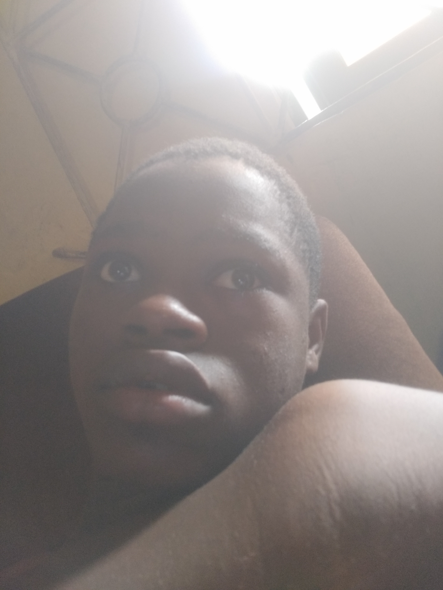 A close-up, slightly overexposed selfie-style photograph shows a dark-skinned individual with short, dark hair. The person's face is prominent, with wide eyes, a broad nose, and slightly parted lips. The individual's left shoulder is visible in the foreground. The background is blurry and poorly lit, but there is what appears to be the ceiling of a room with a ceiling fan. The photograph does not contain any specific details that confirm the location in Shangisha, Nigeria, or any other city. The photograph does not contain any details that describe actions or interactions.