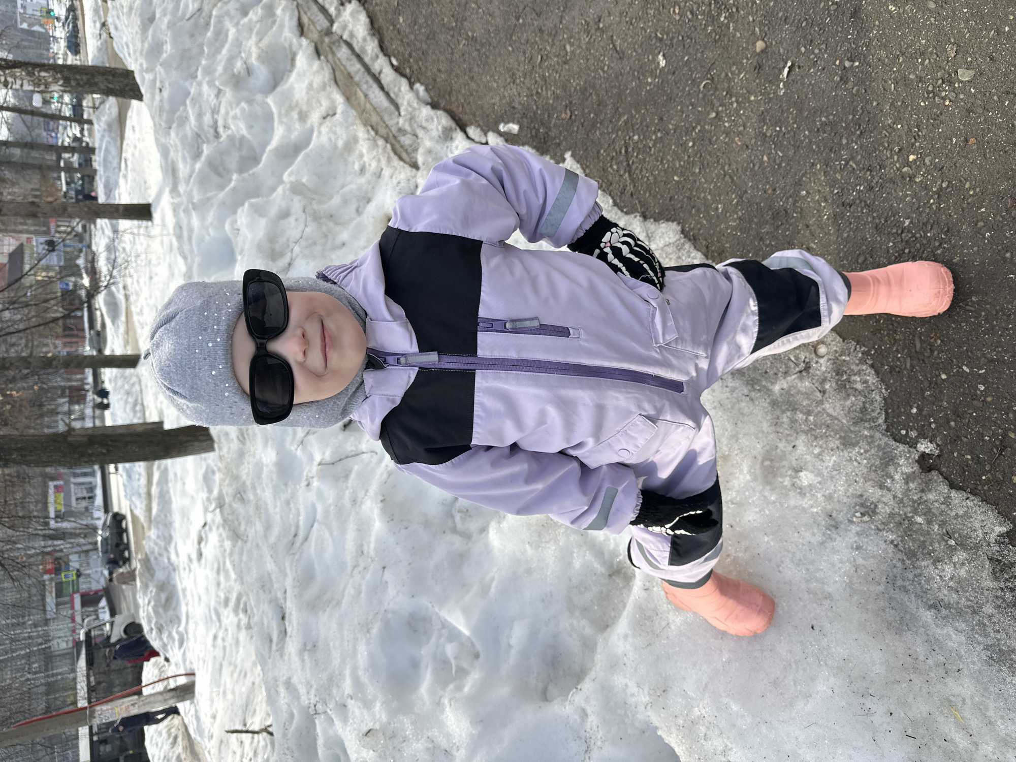 A young child, approximately 3-6 years old, is depicted standing outdoors in Chaikovsky, Russia, on a bright day characterized by melting snow. The child is positioned centrally, wearing a light purple winter overall with black sections on the chest, shoulders, and knees, a grey knitted hat adorned with small reflective embellishments, black rectangular sunglasses, black gloves featuring white skeleton hand patterns, and pink rubber boots. Their left foot is placed on a mound of dirty, melting snow and ice, while their right foot rests on wet asphalt pavement. Their left hand is placed on their hip.
The immediate surroundings consist of patchy snow and exposed pavement. In the midground, large, irregular piles of dirty snow line the edge of what appears to be a sidewalk or path, bordered by bare deciduous trees. The background reveals an urban environment with a street where vehicles are present and parked. Distant buildings, some with commercial facades, are visible, along with additional bare trees and several pedestrians. A red pole is identifiable on the far left of the frame. The scene indicates late winter or early spring conditions.