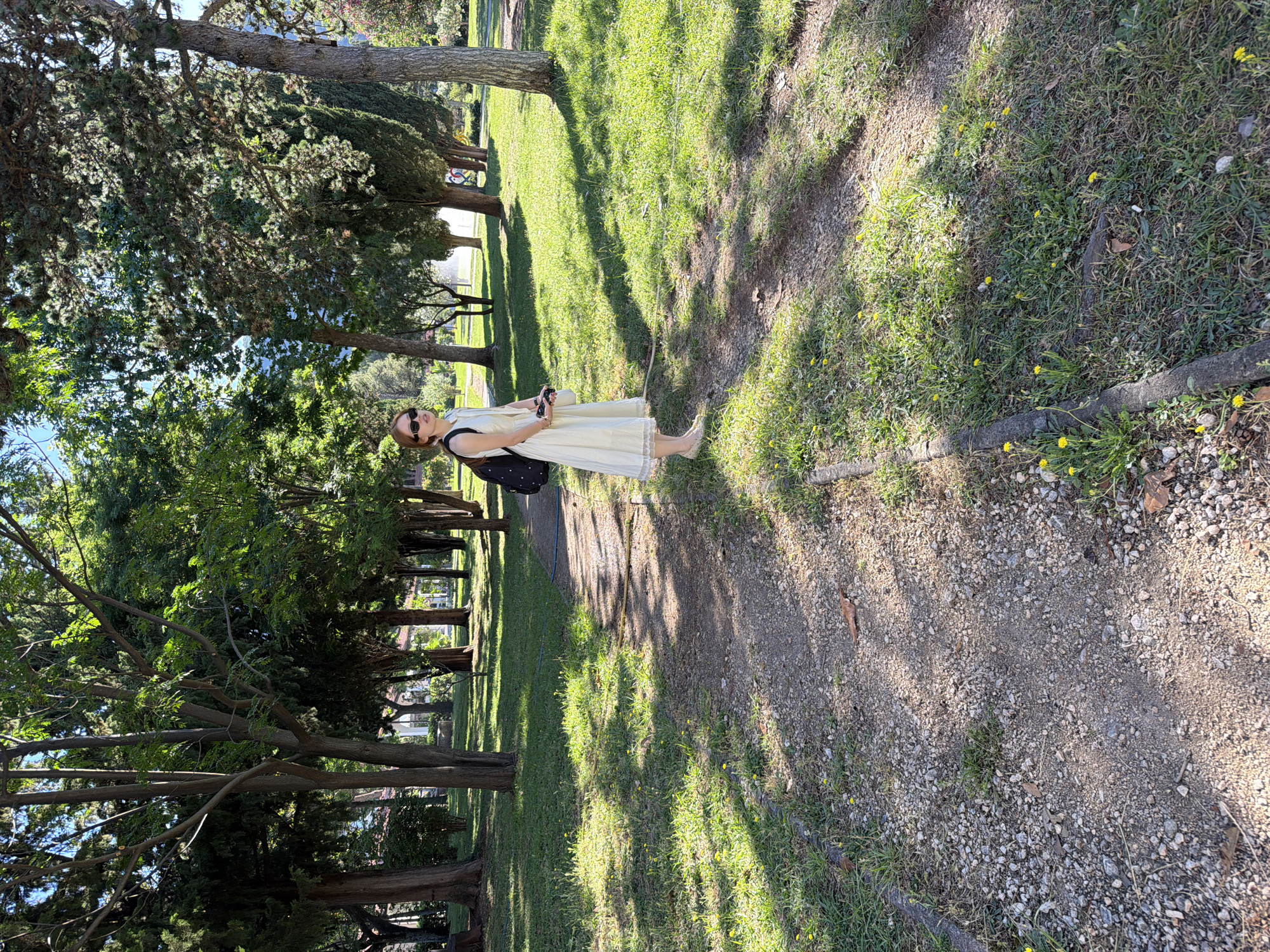 A young woman stands on a gravel path in a park in Budva, Montenegro. She is wearing a cream-colored sleeveless dress with lace trim, beige flats, and sunglasses. A black backpack with white polka dots is slung over her left shoulder. She is looking to her right, with her left hand holding a white object, possibly a phone or camera. The park has lush green grass and numerous trees, some with sparse foliage that allows sunlight to dapple the path and ground. In the background, a fountain is visible, and the trees suggest a hilly or mountainous terrain beyond the park. The path is uneven and edged with grass and small yellow flowers. The overall impression is a sunny day in a well-maintained park.