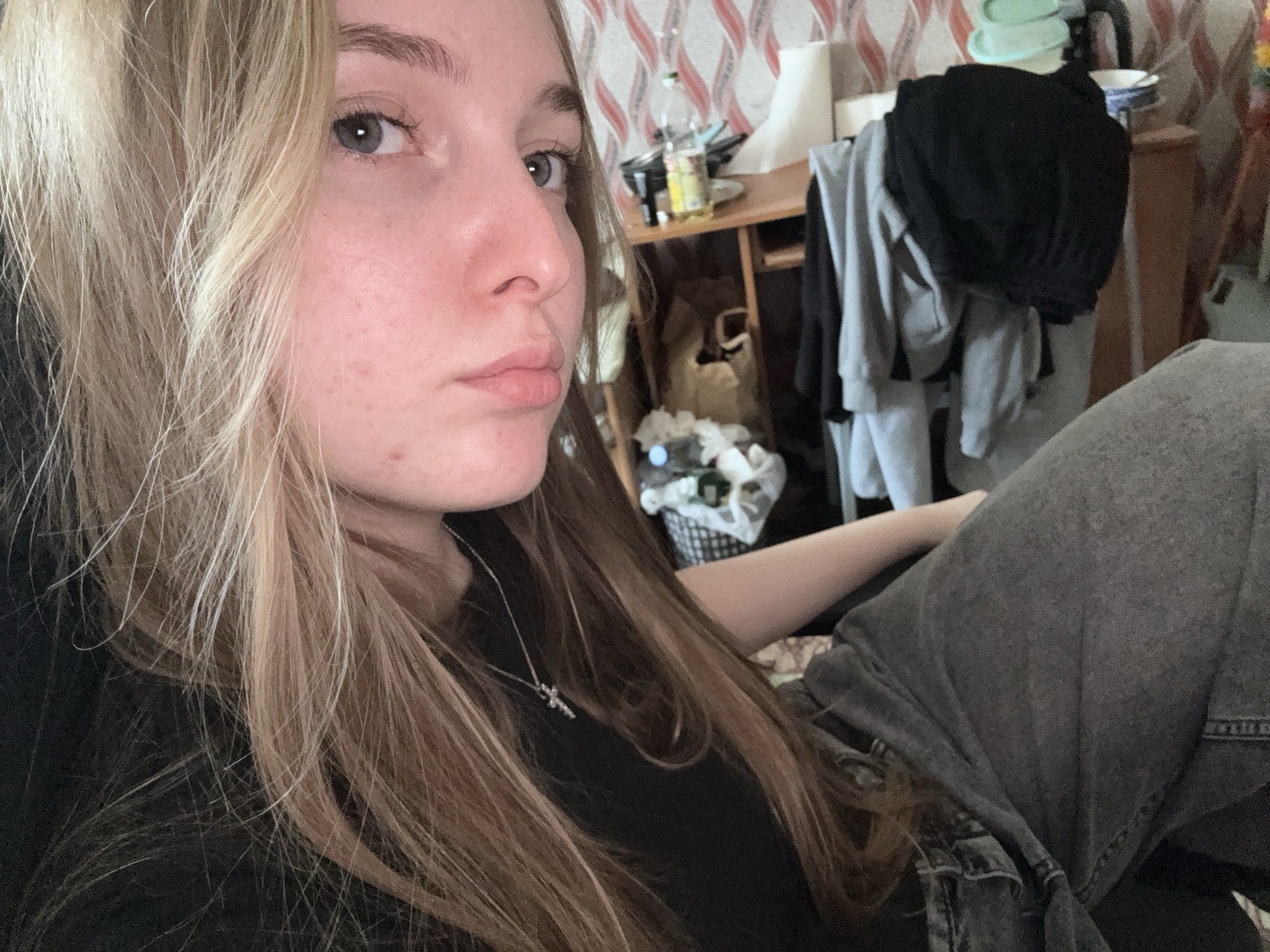 A young woman with light brown and blonde hair is depicted in a selfie. She has fair skin, light eyes, and is wearing a black shirt and a silver necklace with a cross pendant. She is positioned in the foreground with her head turned slightly to the side.
The background reveals a cluttered interior space. There is a wooden shelf cluttered with objects, including a bottle, containers, and miscellaneous items. Clothes are piled on a chair. The wallpaper has a red and white patterned design.
No information from the image reveals anything regarding the city of Perm, Russia.