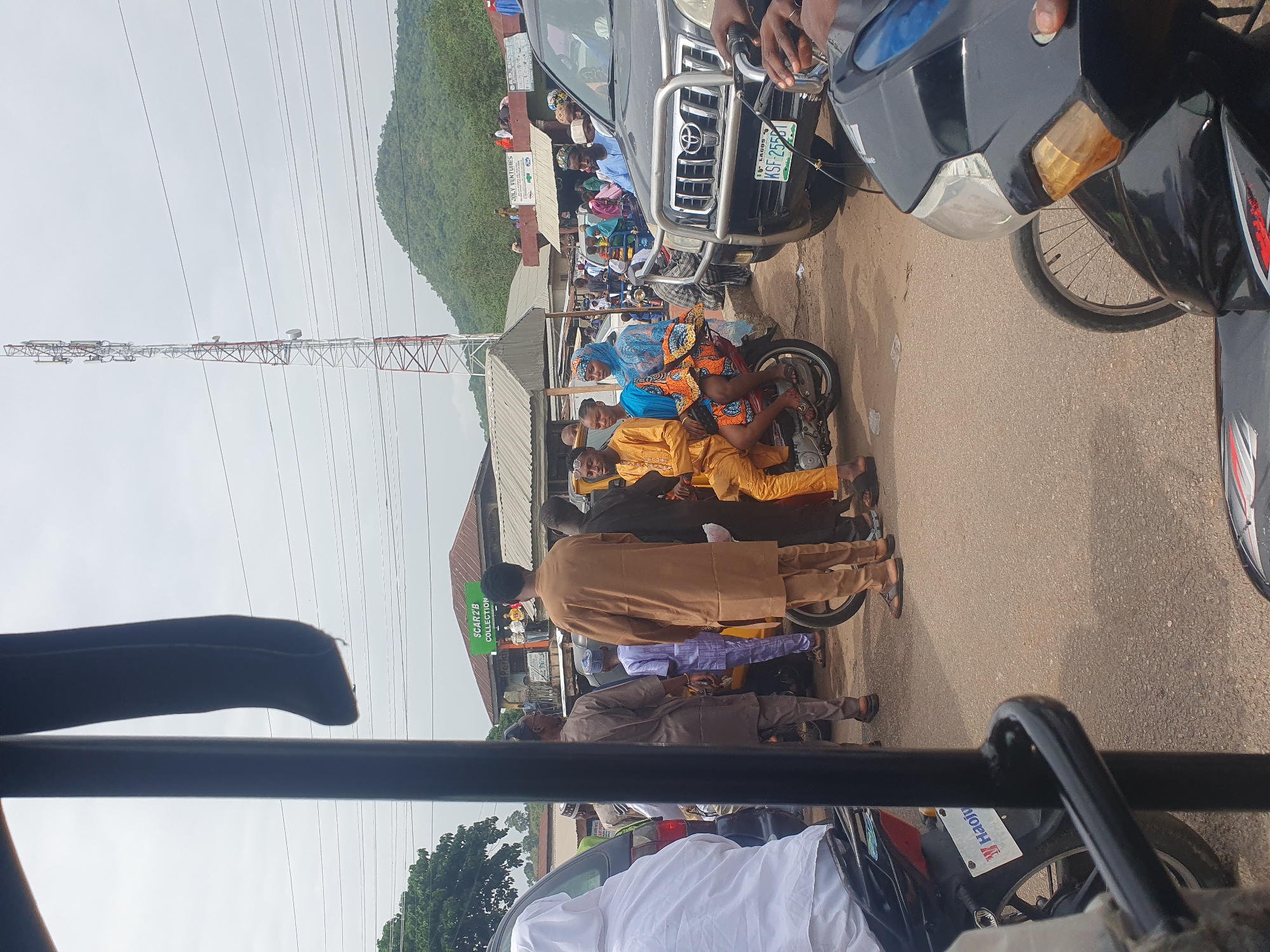 The image depicts a street scene in Lokoja, Nigeria. Several people are present, some on motorcycles and others standing or walking. The individuals on motorcycles appear to be a group, some dressed in traditional clothing. The scene is congested, with people and vehicles in close proximity. A Toyota Prado is visible with license plate KSF-255GT. A power line tower looms in the background. Buildings and trees line the sides of the road, and a hill is visible in the distance. A sign for "SCARZ'B COLLECTION" is present. A motorcycle at the bottom of the photo has the license plate "HAOLI".