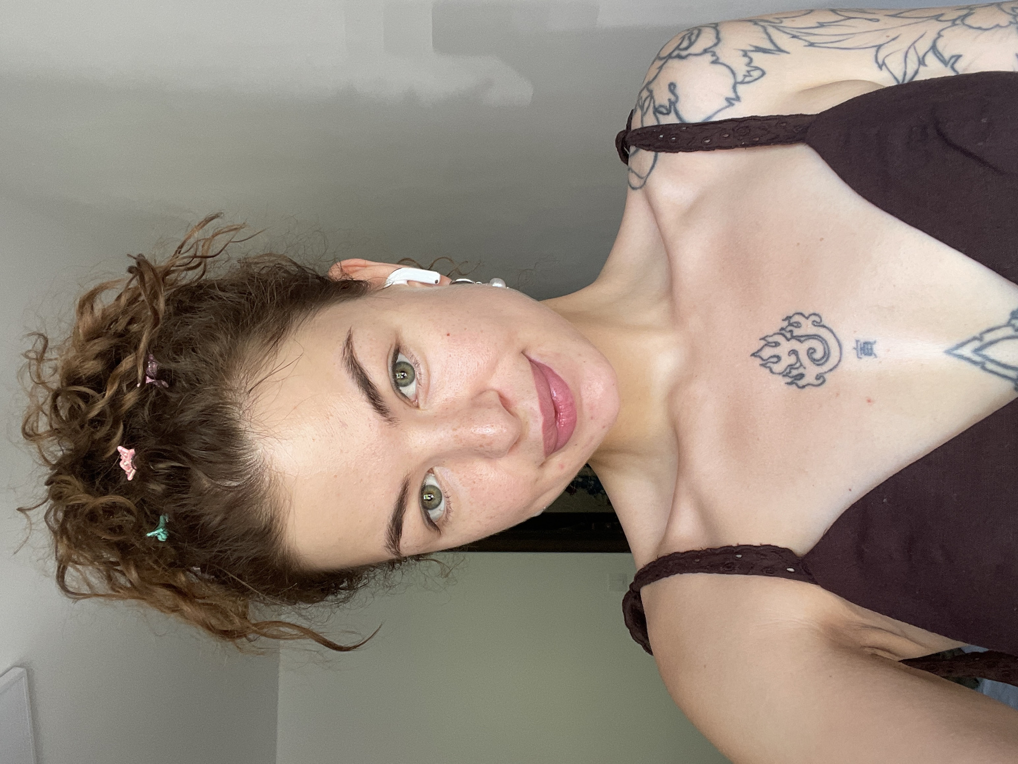 A young woman with light skin and green eyes is pictured from the chest up, looking directly at the camera. Her brown, curly hair is pulled up in a messy bun, adorned with small butterfly clips in pink and teal. She is wearing a dark brown top with braided straps. A white earbud is in her right ear, and she has a silver earring in her left ear. She has freckles across her nose and cheeks, and her lips are painted a soft pink. A tattoo of stylized flames is visible on her chest, above another smaller tattoo. A large floral tattoo is visible on her left shoulder and arm. The background is a plain, light gray wall. The image appears to be taken indoors.