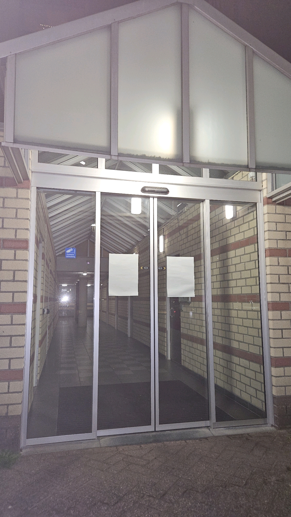 The image shows the exterior of an automatically opening sliding glass door entrance to a building. The building's facade is constructed of red brick with yellow accents. Above the glass doors is a frosted glass canopy with metal framing. Inside the building, a well-lit corridor with a checkered floor extends into the distance. There are two blank white signs attached to the glass doors. A blue sign with white text and an icon of a security camera is visible on the left side of the interior corridor, indicating "Camera bewaking," which translates to "Camera surveillance" in Dutch. The scene appears to be located at the entrance to the Galderse Heide sports park in Breda, Netherlands, based on the "Camera bewaking" sign.