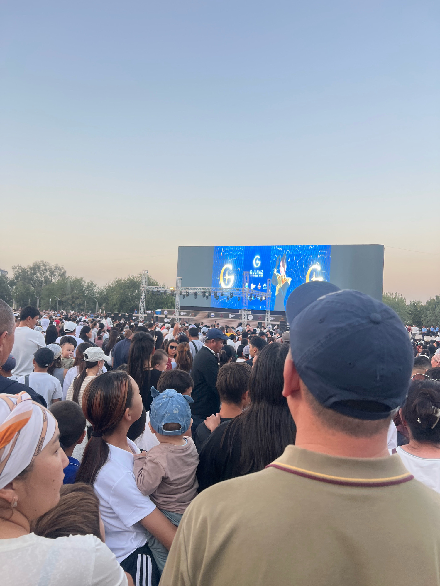 The image depicts a large gathering of people outdoors, likely at an event in Osh City, Kyrgyzstan. A large screen is visible in the background, displaying a logo that includes the letter "G" and the word "GULNAZ." Several individuals are observing the screen, suggesting a performance or presentation is taking place. The crowd is diverse, with adults and children present. The environment appears to be a park or open public space, with trees and a clear sky in the background. The time of day seems to be late afternoon or early evening, given the lighting conditions. The overall scene suggests a community event or festival.