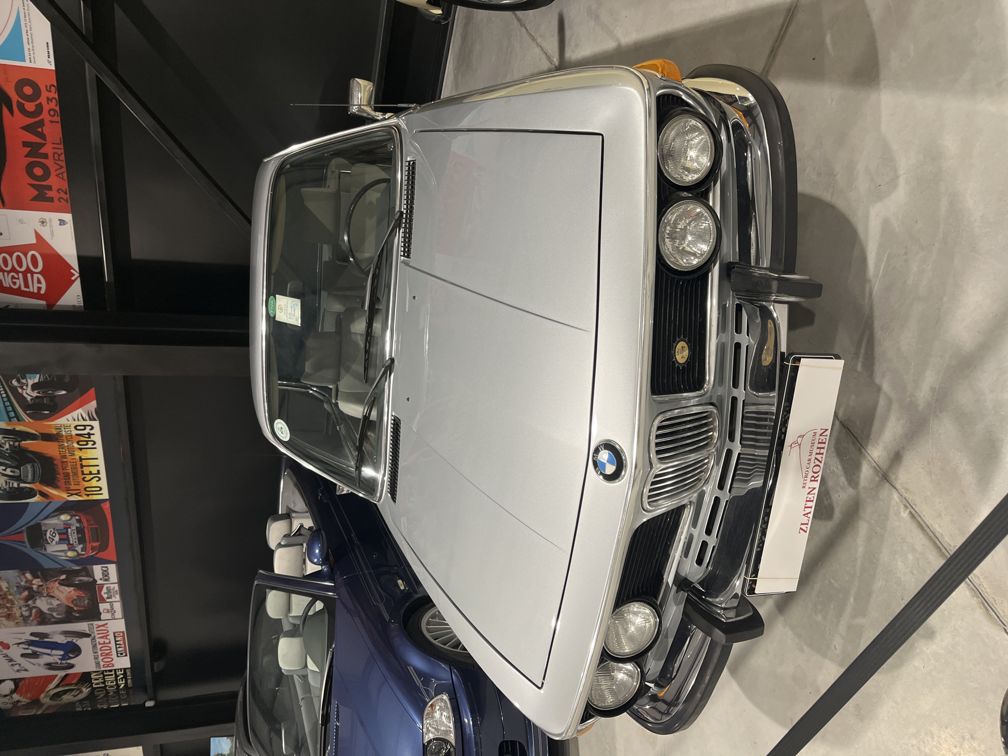 The image shows a silver BMW coupe with the emblem of the "Zlaten Rozhen" Retro Car Museum affixed to the front. The museum is located in Kapatovo, Bulgaria. The car has a classic design with four headlights and a chrome grille. To the left is a blue car. On the left, hung against a black wall, are posters depicting vintage automobile races. These include the Monaco Grand Prix of April 22, 1935, the Bordeaux Automobile Motocycliste race, and the 10 Sett 1949 Grand Prix.
