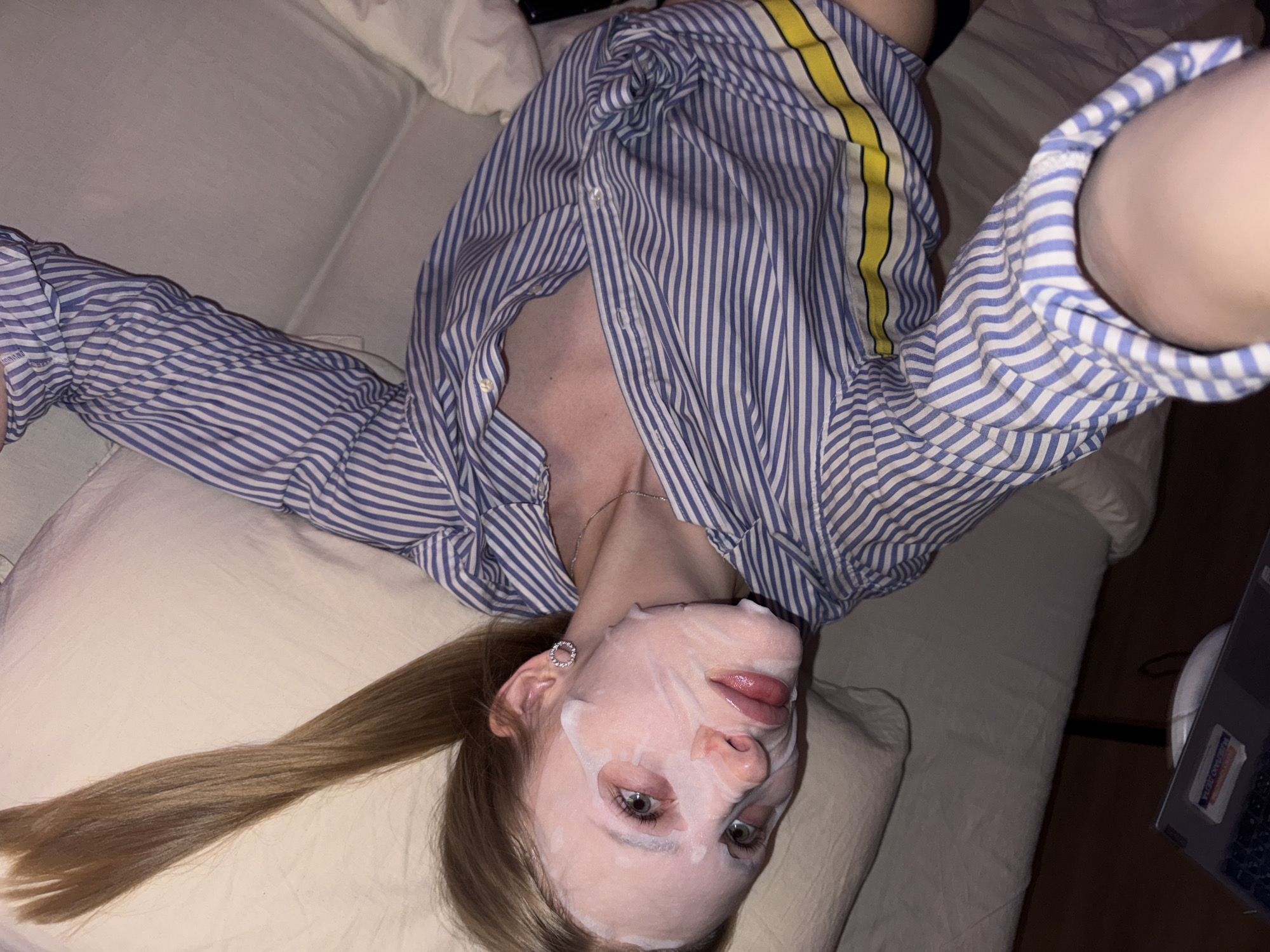 A young individual is lying on a light-colored couch, positioned on their back. They are wearing a blue and white striped shirt with a yellow accent stripe on the shoulder and have a facial mask applied. The individual has long blonde hair which rests on a pillow. A laptop is visible in the background to the right. No information indicates that the image was taken in Perm, Russia.