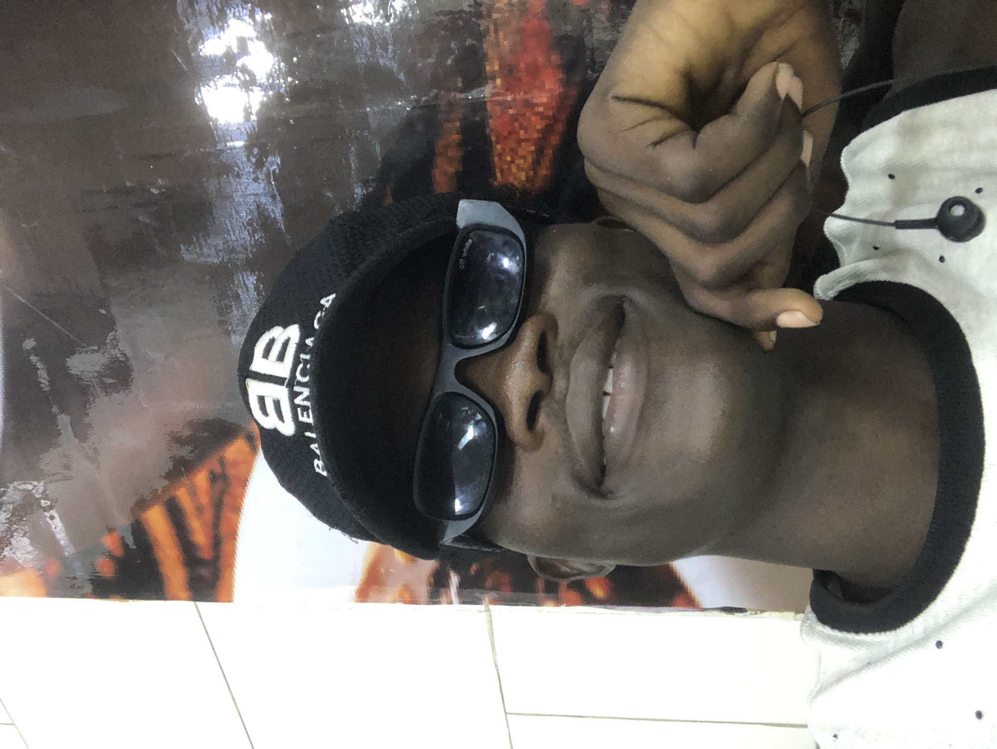 A young Black male is captured in a selfie, wearing a black Balenciaga baseball cap and black sunglasses. He is leaning his head on his right hand, which is resting on his cheek. His mouth is open in a smile, revealing his teeth. He is wearing a white t-shirt with black trim around the collar. A black earphone cord is visible hanging from his shirt. The background appears to be a wall with a large, glossy mural or advertisement, featuring abstract shapes and colors, possibly in a commercial or public space. The overall lighting suggests an indoor setting.
