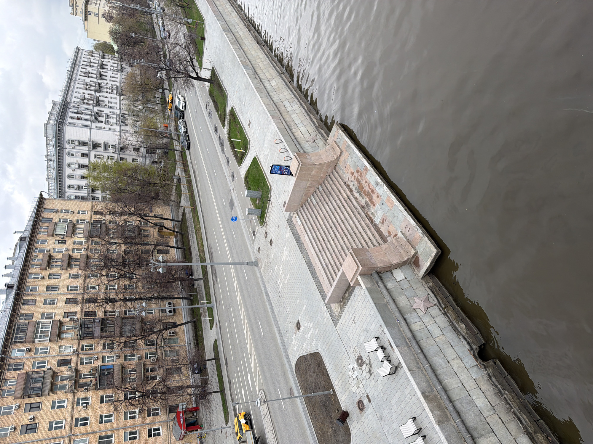 This high-angle, overhead shot captures a section of a riverside embankment, a multi-lane road, and urban buildings under an overcast sky. The scene is presented from a tilted perspective, looking down from a significant height, typical of an aerial or drone view.
On the right, a wide, dark-grey river flows, showing subtle ripples on its surface. It is bordered by a robust stone embankment with multiple levels. A broad flight of stone stairs, constructed from lighter-colored stone, descends from the upper promenade towards the water. On the upper promenade, several white benches are arranged, and a digital information screen displays blue and white graphics, possibly related to a bike-sharing service, accompanied by bike racks. A distinctive star-shaped stone ornament is embedded in the lower part of the embankment wall near the water's edge.
Parallel to the embankment runs a multi-lane asphalt road, clearly marked with white lines. Several parked cars, including a yellow vehicle, are visible. Tall lamp posts line the road and embankment, and patches of green grass and bare trees are interspersed throughout the urban landscape, suggesting early spring or late autumn. A circular blue sign with a white bicycle icon indicates a bicycle path.
On the left side of the image, two prominent buildings dominate the background. The closer structure is a multi-story, light brown brick building with numerous windows, balconies, and external air conditioning units. Further down the street stands a more architecturally elaborate, lighter-colored building, characterized by its grand scale, ornate details, arched windows, and multiple balconies, indicative of Stalinist-era architecture common in Moscow.
The overall impression is one of a quiet urban scene, likely captured during daytime on a cloudy day in Moscow, Russia, given the architectural style and urban infrastructure elements. No explicit human activity is visible, making the scene appear still and composed.