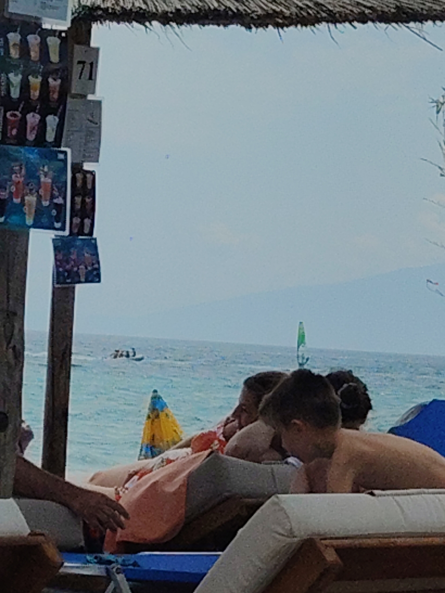 The image depicts a beach scene, likely in a coastal area. In the foreground, several people are seen relaxing on lounge chairs under a thatched roof structure. One person's arm is visible, resting on the chair. Behind them, the turquoise sea stretches out to the horizon, with a faint outline of land in the distance. A few boats or watercraft are visible on the water. The sky is a pale blue, suggesting a clear day. There are some signs with images of drinks attached to a wooden pole on the left side of the image. The overall atmosphere is one of leisure and vacation. The mention of Ωραιόκαστρο, Greece, suggests a specific geographic location, though it is not visually evident from the image itself.