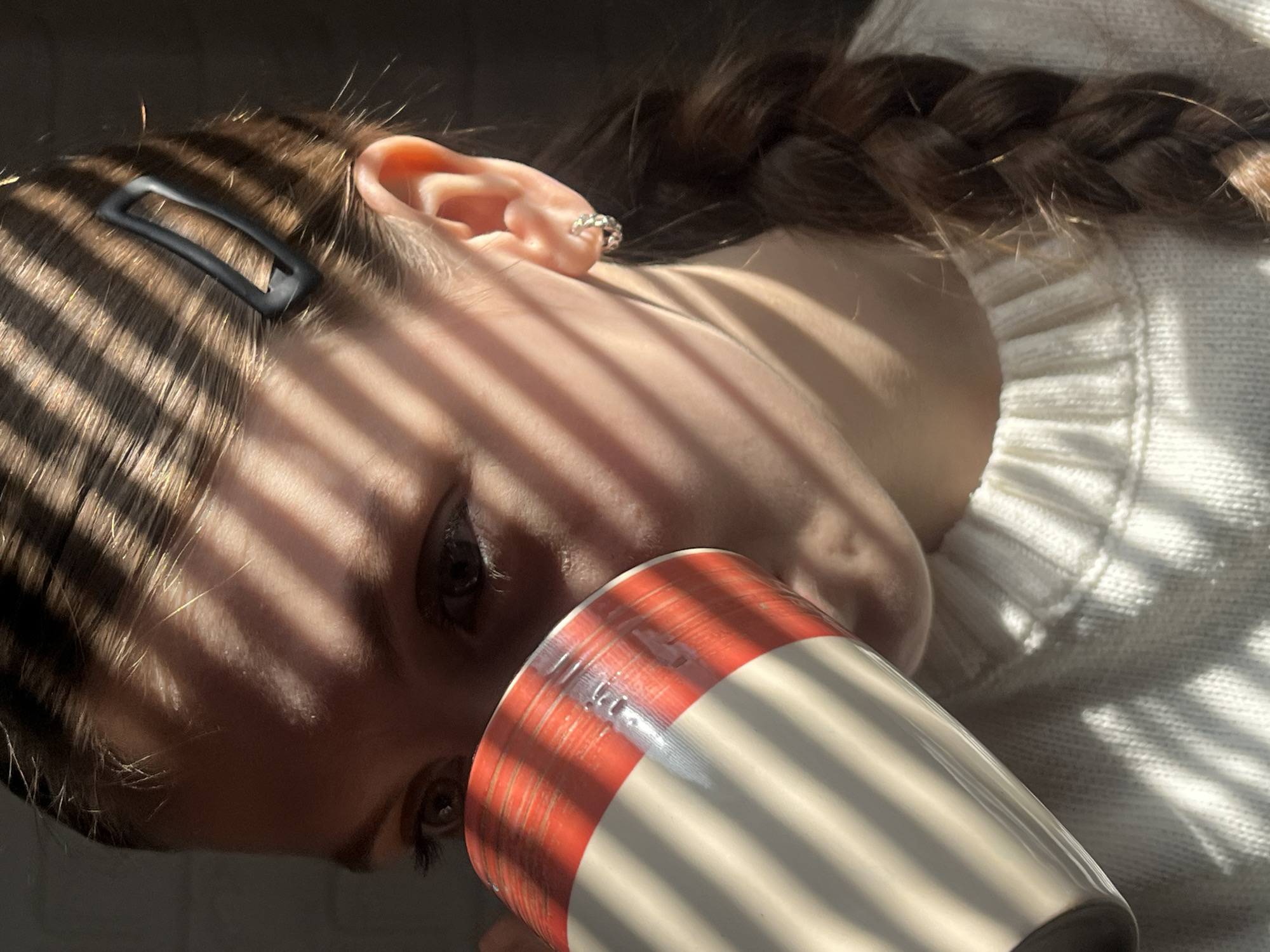 A person is seen from a close, high-angle perspective, holding a red and white striped mug. The person has a braid, a hair clip, and an earring in their ear. Light from a window casts linear shadows across their face and clothing. They are wearing a white sweater. The city of Фоки, Russia, cannot be verified as part of the image's location.