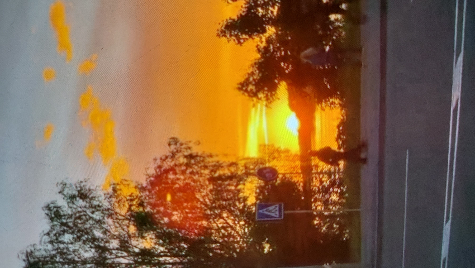 A sunset is captured over a body of water, with the sun appearing as a bright, glowing orb. Silhouetted trees line the shore, framing the vibrant orange and yellow hues of the sky. In the foreground, a paved sidewalk runs parallel to a road, with two indistinct figures walking on the sidewalk. One figure is closer to the left side of the image and appears to be walking away from the viewer. The other figure, to the right, is also walking away. A pedestrian crossing sign is visible on the left side of the sidewalk. The overall scene suggests a public space near water, likely a park or promenade, during twilight.