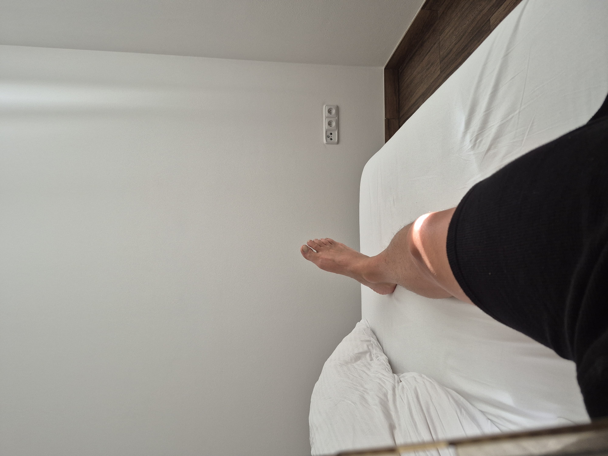 The image depicts a person's legs lying on a white-sheeted bed in a room. The bed is positioned in the lower half of the frame, with a white pillow visible on the left. The person is wearing black shorts. A shaft of sunlight illuminates the person's thigh. The background consists of a plain, light-colored wall with three electrical outlets mounted on it. The wall meets a wooden floor at the bottom right corner. The overall scene suggests a domestic or hotel room setting, likely in Poděbrady, Czechia, given the presence of European-style electrical outlets and the context provided.