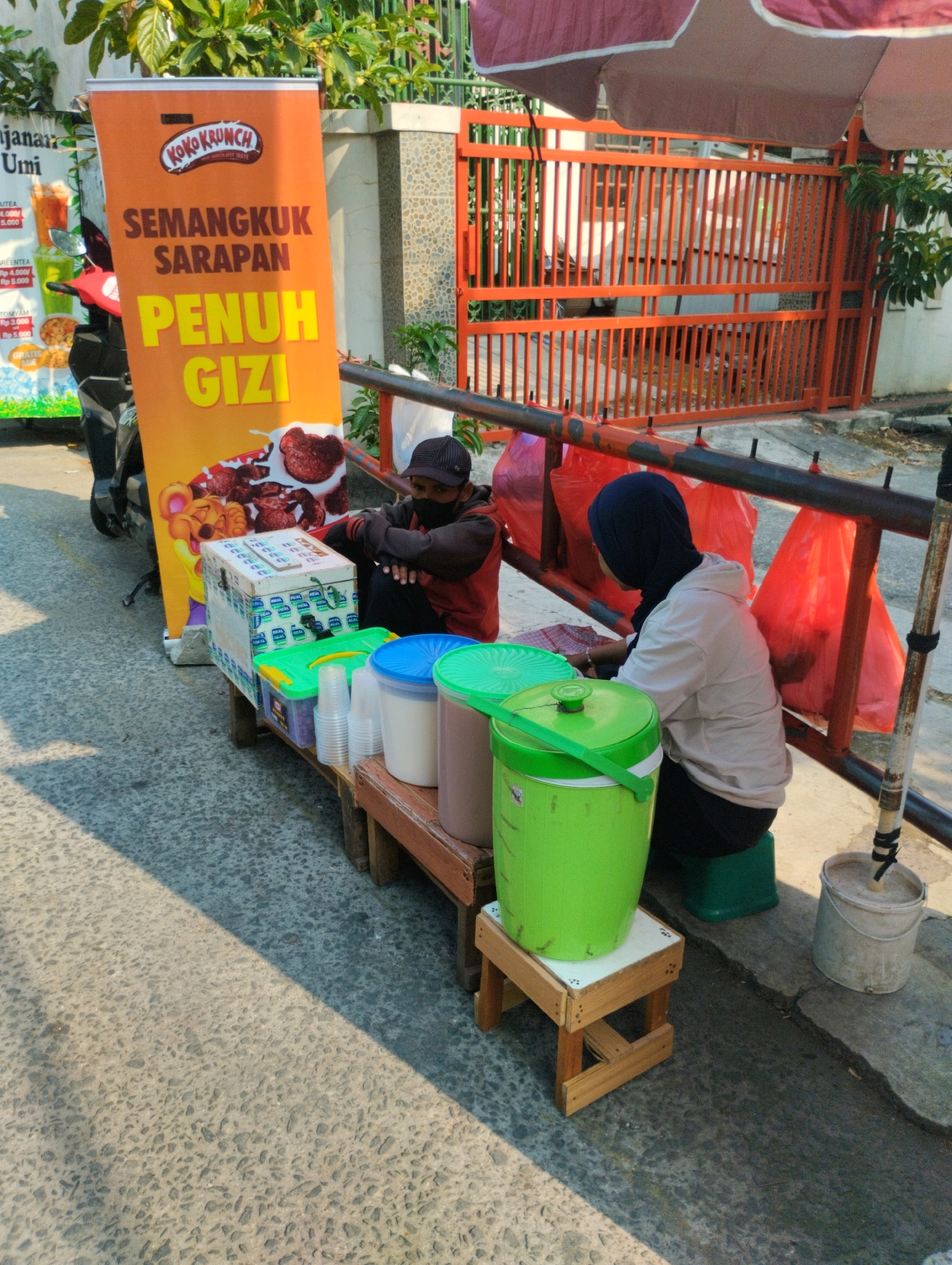 A street food stall operates on a sun-drenched asphalt road in front of a fenced orange gate, with lush green foliage visible above. The stall features a prominent orange banner with "KOKOKRUNCH" and "SEMANGKUK SARAPAN PENUH GIZI" (A bowl of nutritious breakfast) printed on it. A man wearing a black cap and a face mask sits behind a display of plastic containers and cups, suggesting beverages or food items are being served. To his right, a person wearing a hijab and a light-colored hoodie sits with their back to the camera, facing away from the stall. Red plastic bags hang from a metal railing behind the stall. The environment appears to be urban, potentially a residential area or a commercial street within Bekasi, Indonesia, given the context provided.