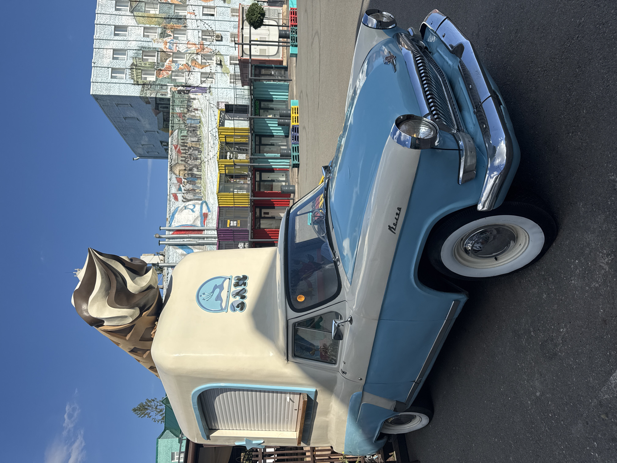 A vintage blue and white truck, modified into an ice cream stand with a large ice cream cone sculpture on top, is parked in an outdoor area. The truck has a closed roll-up window on the side and a wooden counter. A logo featuring a deer and the Cyrillic word "BAR" is visible on the side of the truck's enclosure. In the background, a building is adorned with a large mural depicting various scenes and figures. Several colorful shipping containers, also serving as storefronts, are arranged in front of the muraled building, some with yellow string lights overhead. The ground is paved asphalt, and the sky is clear and blue. The scene appears to be in the city of Ufa, Russia, specifically at the Ufa Arena parking lot, which has been transformed into a pedestrian zone with the shipping container shops. There are no people clearly visible in the immediate vicinity of the truck or the shipping containers.