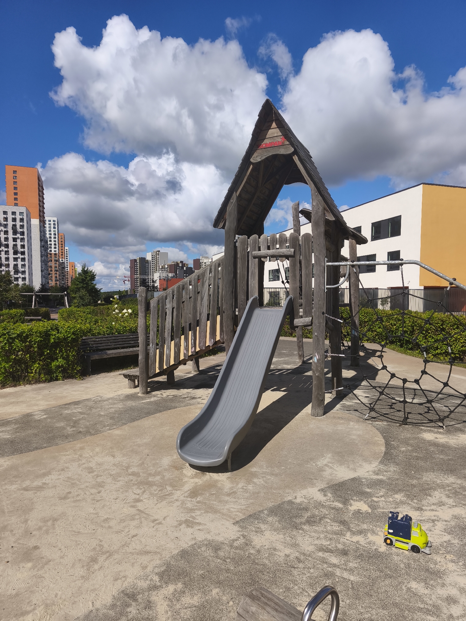 The image shows a children's playground in a modern urban residential area, possibly in Moscow, Russia, indicated by the architectural style of the surrounding high-rise buildings. The playground features a wooden play structure with a grey slide, climbing ropes, and wooden planking. In the foreground, a small yellow and blue toy truck rests on the sandy ground. Behind the playground, a dense green hedge lines the area, with a wooden bench visible within the greenery. The background is dominated by several tall apartment buildings of varying designs and colors, under a bright blue sky with fluffy white clouds. The overall scene depicts a well-maintained public space within a newly developed urban environment.