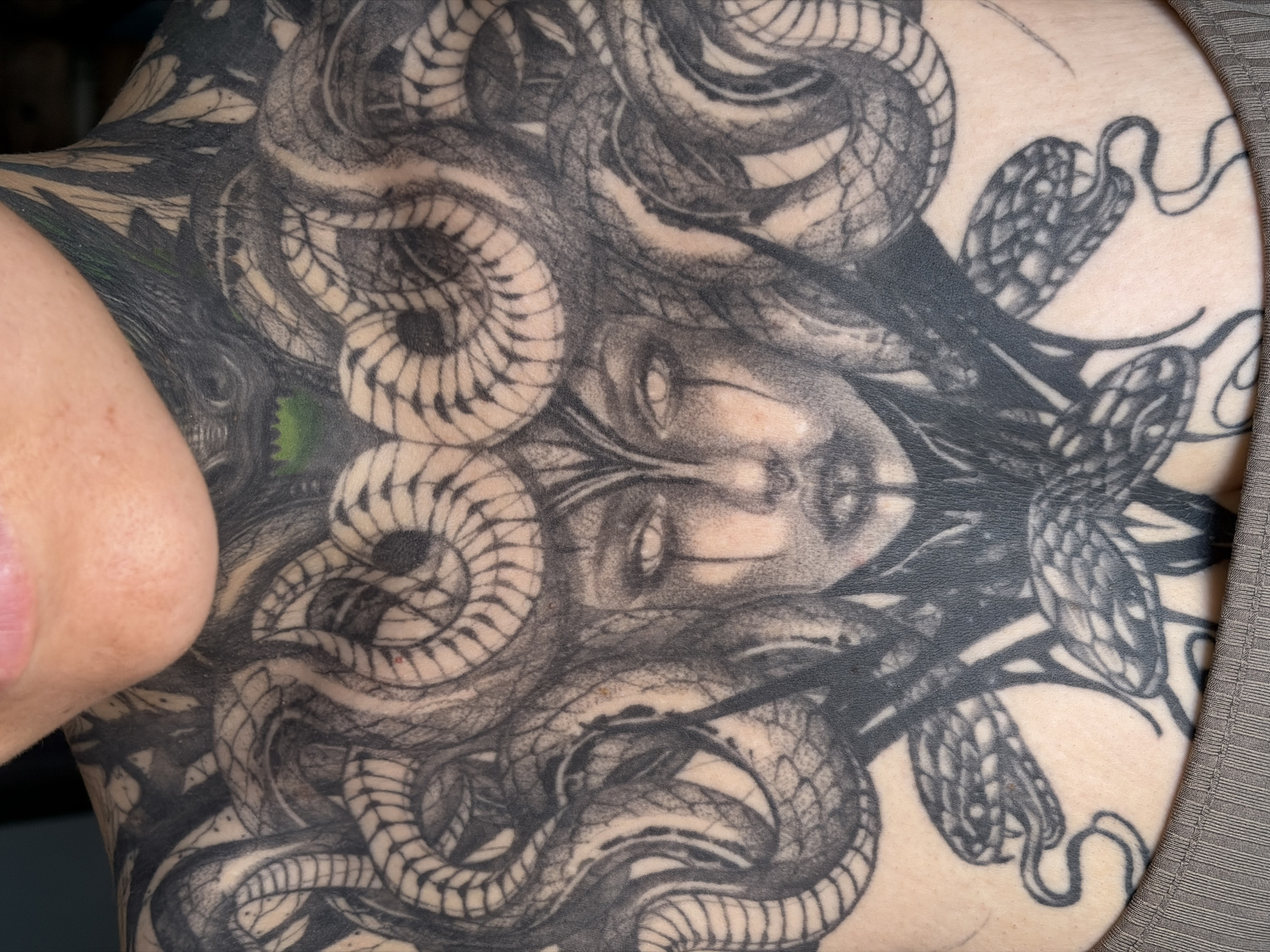 The image shows a close-up of a tattoo on a person's chest and neck. The tattoo depicts Medusa with snakes for hair. The detail and shading of the tattoo are intricate, with the snakes' scales and Medusa's facial features clearly visible. Some of the snakes are coiled around Medusa's head, while others are positioned lower down on the chest. The person's skin tone is fair. The upper lip and chin of the person are visible at the top of the frame, and the top of a gray ribbed garment is visible at the bottom. There is no indication of the city of Chaikovsky, Russia in the image, or any information that would place it there.